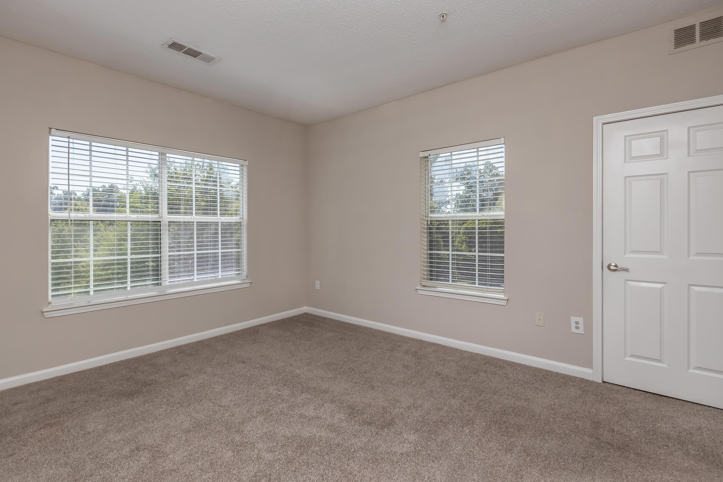 A vacant room with beige walls and light brown carpet. Two windows with white blinds allow natural light in, offering a view of greenery outside. A white door leads to another space, and there are no furnishings, creating a spacious, neutral atmosphere.