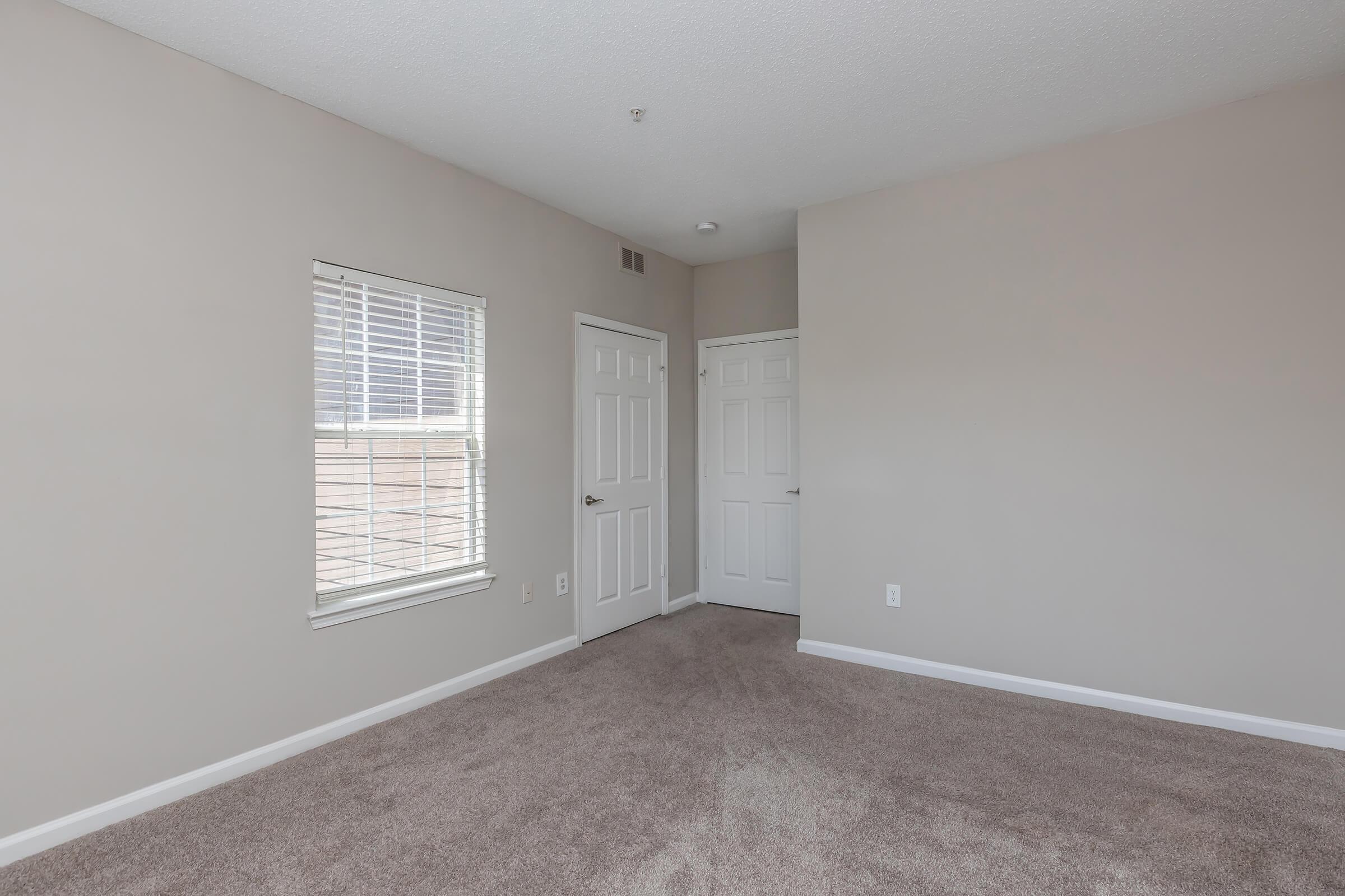 A light-filled, neutral-colored room featuring beige walls and a plush carpet. There is a window with white blinds on one wall and two closed doors on the opposite wall, indicating access to additional spaces. The overall atmosphere is spacious and inviting, suitable for various uses.