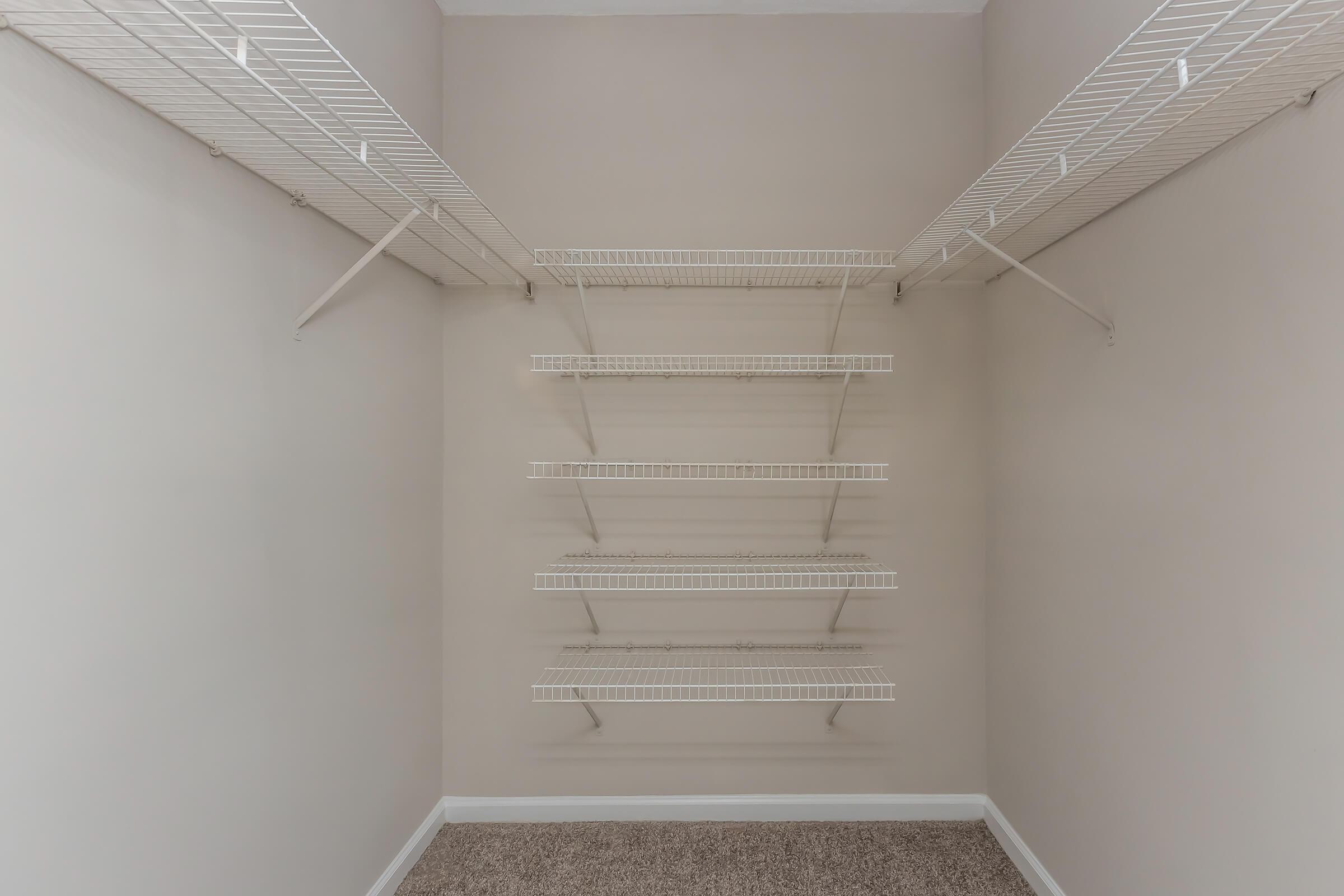 An empty closet with white wire shelving on both sides. The walls are painted a neutral color, and the floor is covered with carpet. The shelving is evenly spaced, providing multiple levels for storage, but there are no items displayed on the shelves.