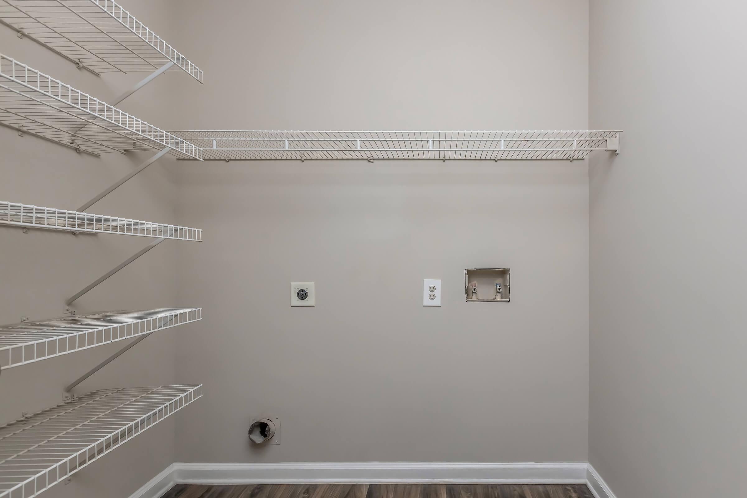 A spacious, empty closet featuring white wire shelving on two walls. The walls are painted a neutral beige, and the floor is wood-like laminate. There is an electrical outlet and a small recessed opening on one wall, suggesting it may be designed for appliances.