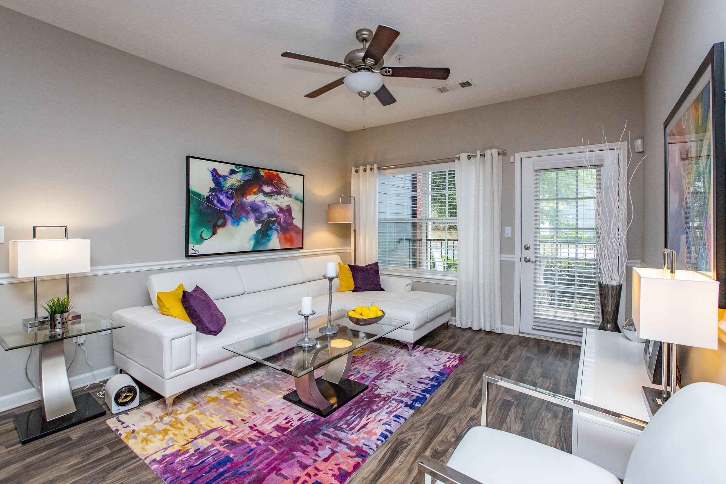 A modern living room featuring a white leather sofa, a glass coffee table with a bowl of yellow fruit, and a colorful area rug. The walls are painted a soft gray, adorned with an abstract painting, with large windows draped in white curtains and a ceiling fan overhead. Bright and inviting decor creates a cozy atmosphere.