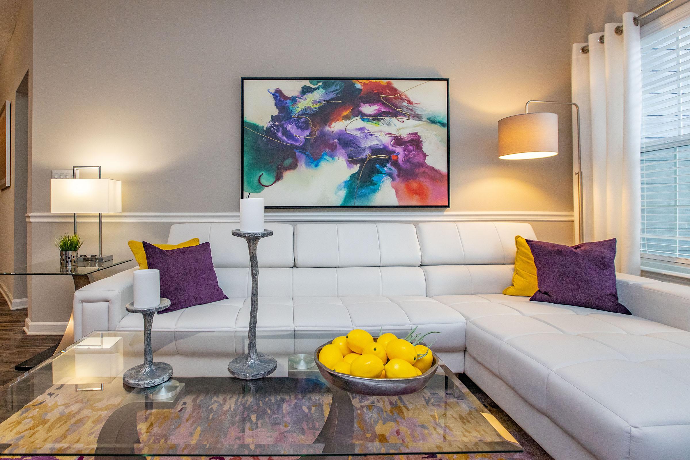 A modern living room featuring a white sectional sofa adorned with purple cushions, a glass coffee table with a bowl of bright yellow lemons, and elegant candle holders. A vibrant abstract painting hangs on the wall, complemented by stylish lighting fixtures that create a warm ambiance.