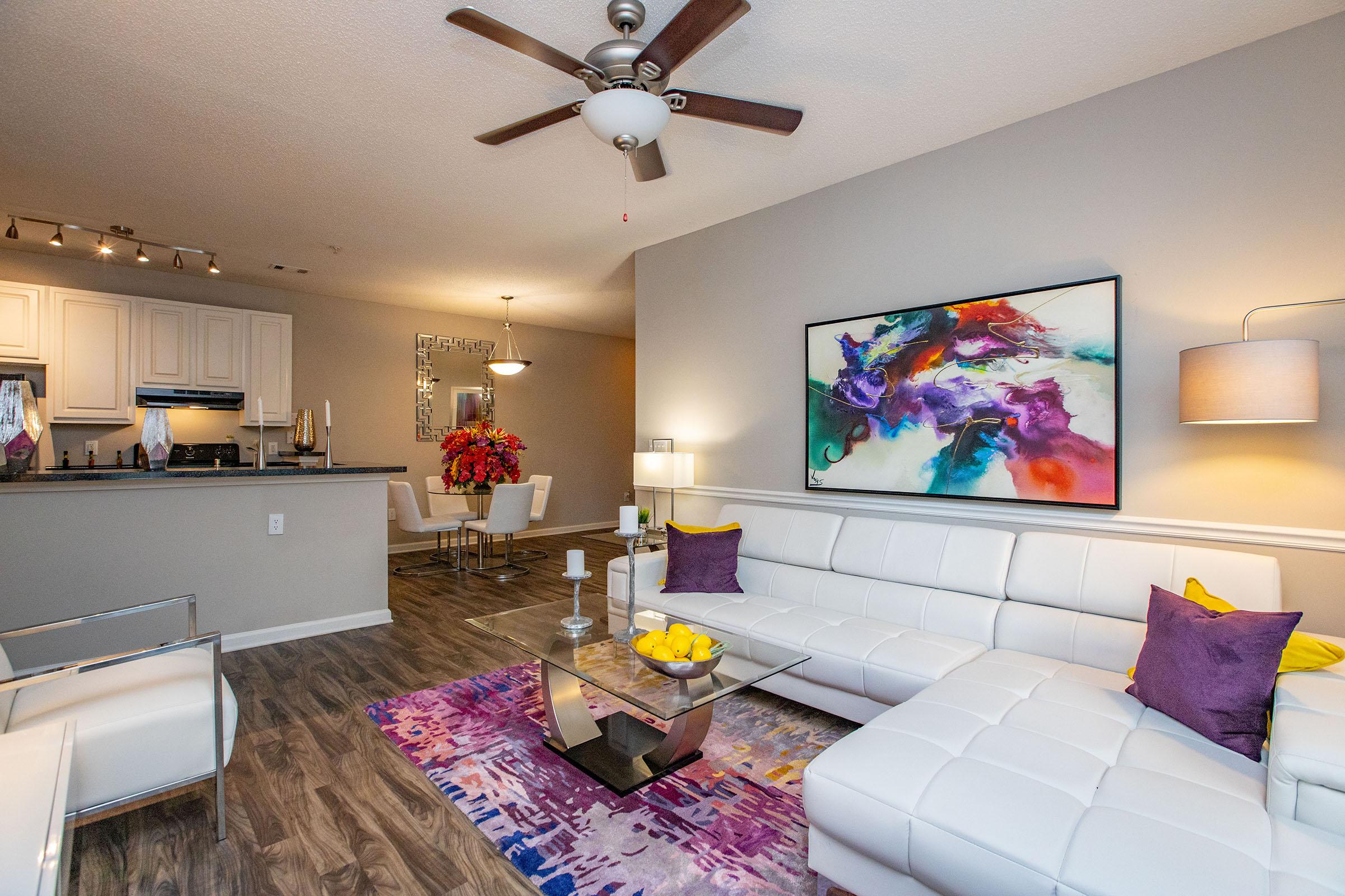 A modern living room featuring a white sectional sofa with colorful accent pillows, a coffee table with a glass top, and decorative items. The room has a ceiling fan and a vibrant wall art piece. In the background, there is a dining area with a round table and chairs, complemented by stylish decor.