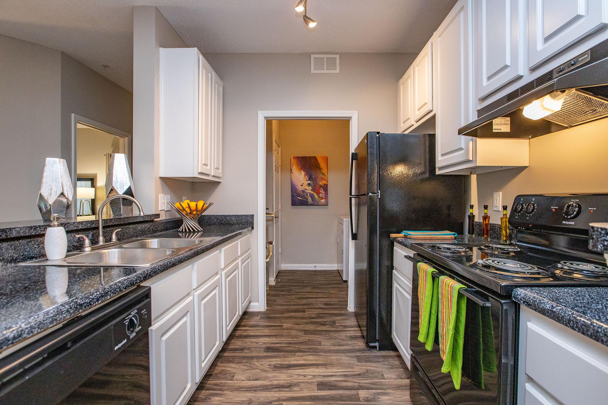 A modern kitchen featuring white cabinetry, black appliances, and a granite countertop. There’s a double sink, a stove, and an oven, along with vibrant green kitchen towels hanging. In the background, a doorway leads to another room, and there are decorative items on the counter, including a bowl of bananas.