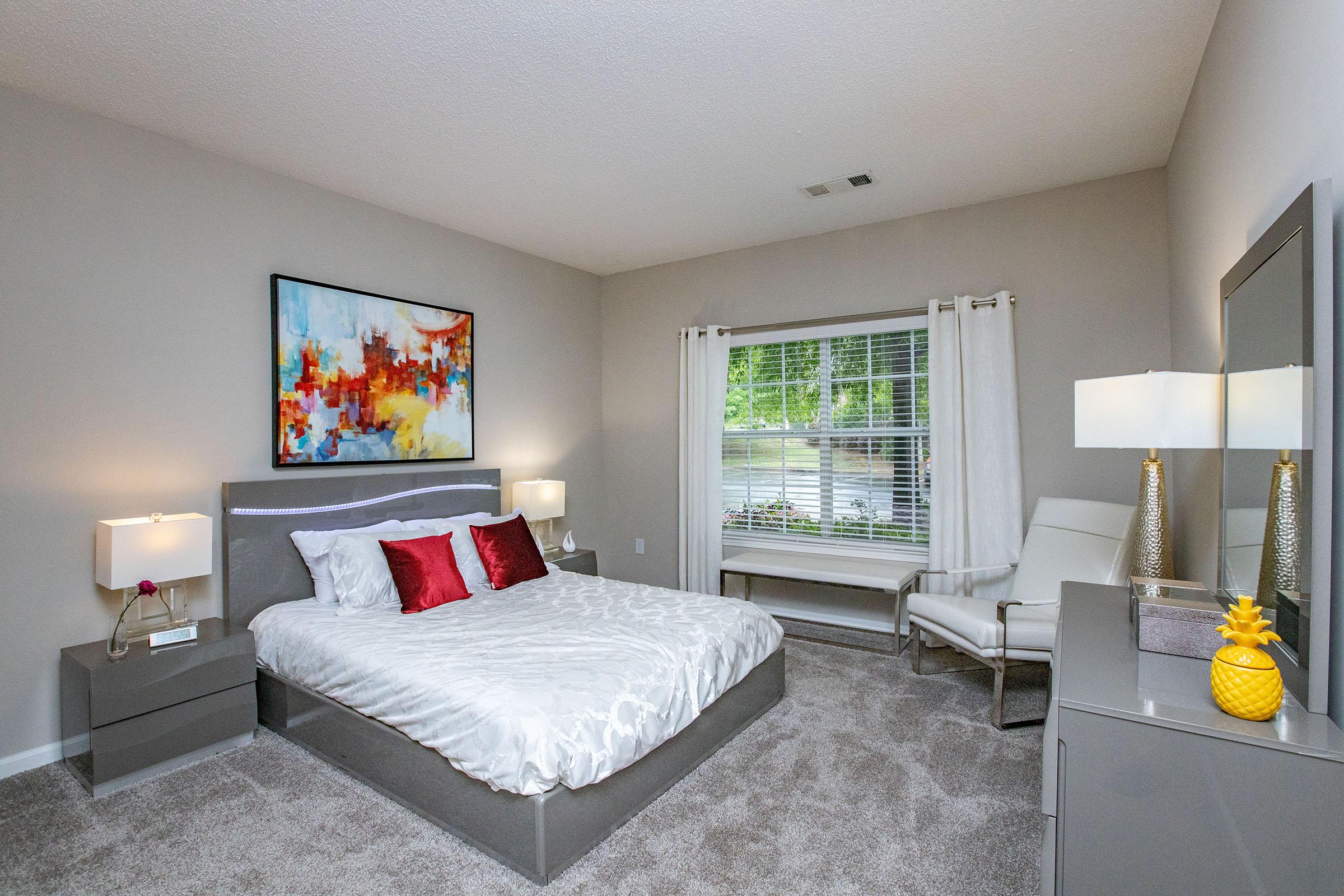 A modern bedroom featuring a large bed with white linens and red accent pillows. There are two bedside lamps on gray nightstands, a comfy chair near the window, and a stylish dresser with a decorative pineapple. Natural light fills the space, complemented by soft, neutral wall colors and artwork above the bed.