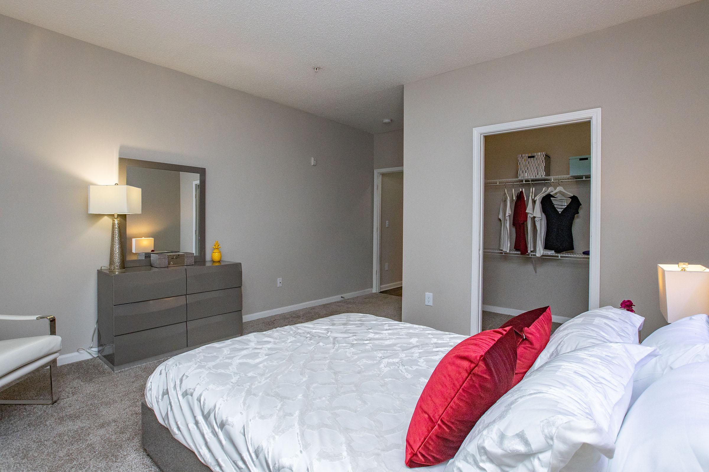 A spacious bedroom featuring a large bed with white bedding and a red decorative pillow. A gray dresser is positioned near a lamp, with a mirror above it. The room has neutral-colored walls and carpet, and a closet with clothing visible in the background. Soft lighting creates a cozy atmosphere.