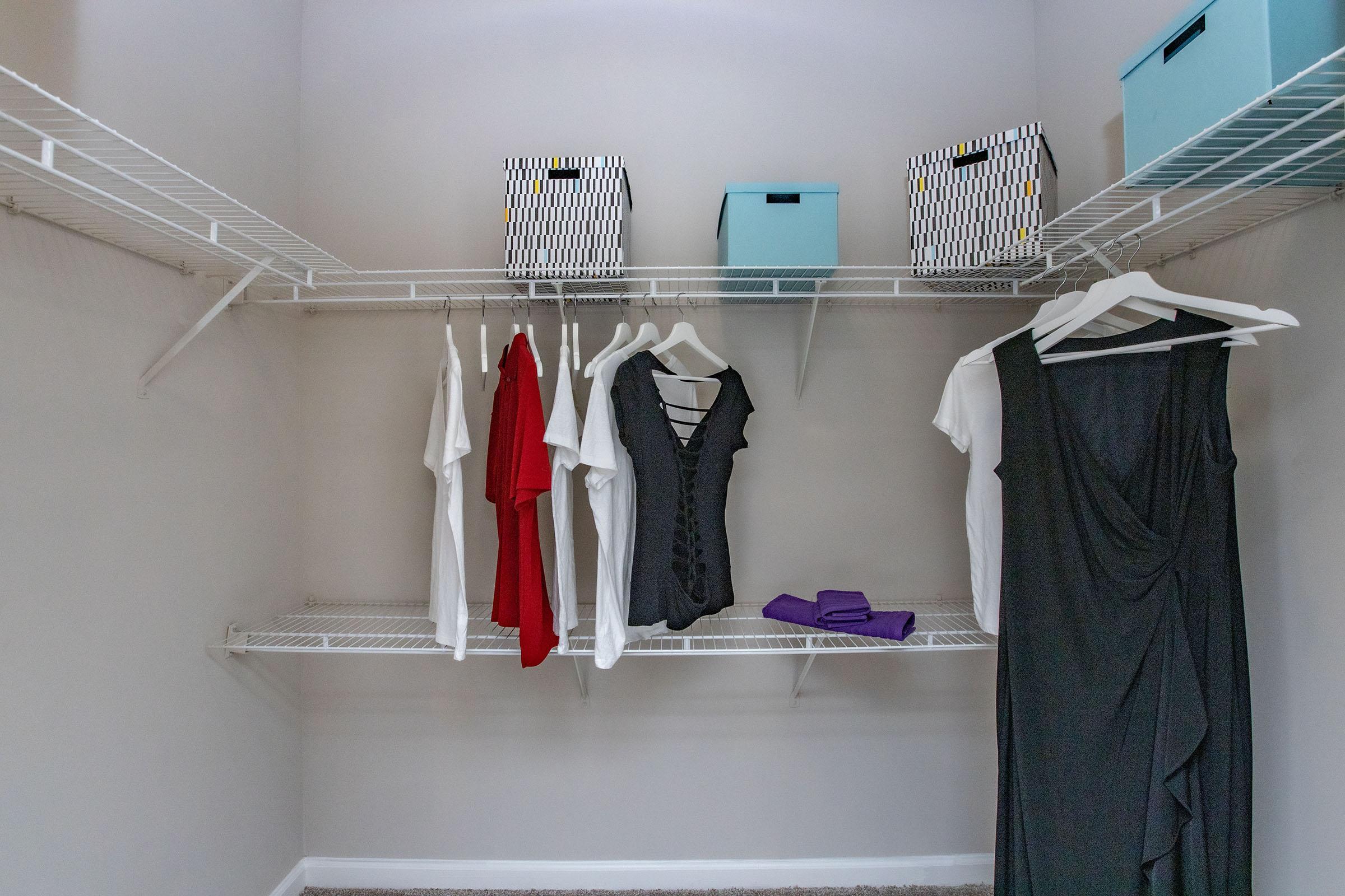 A neatly organized closet featuring several hanging shirts in white and red, a black dress, and a folded purple towel. There are also decorative storage boxes in blue with a black and white geometric pattern on the upper shelves. The walls are light-colored and the floor is carpeted.