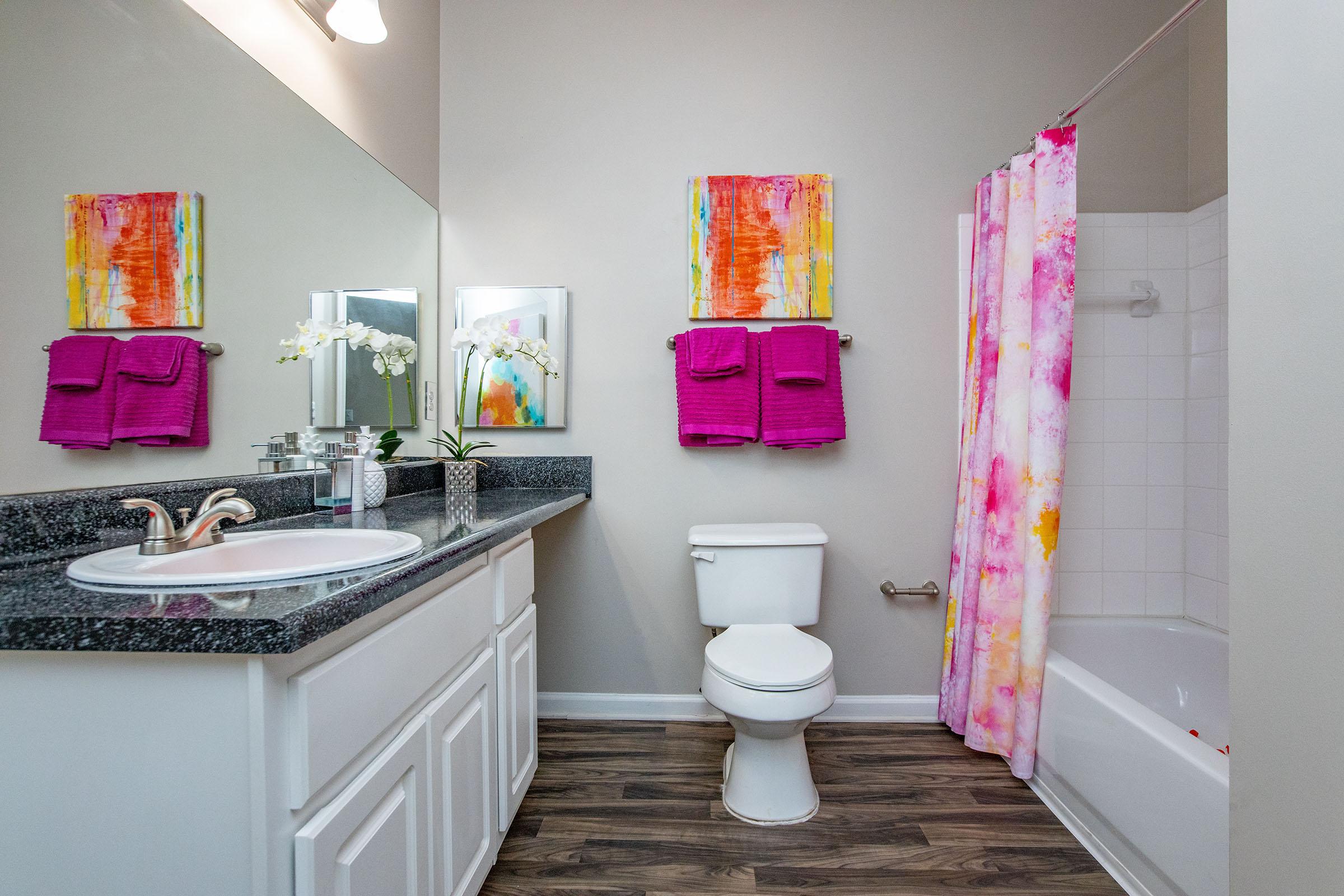 A modern bathroom featuring a white toilet and a bathtub with a shower curtain in vibrant pink and orange tones. The vanity has a dark countertop with a round sink, and there are two colorful abstract art pieces on the walls. Pink towels hang neatly, and there are decorative touches like a plant and flowers.