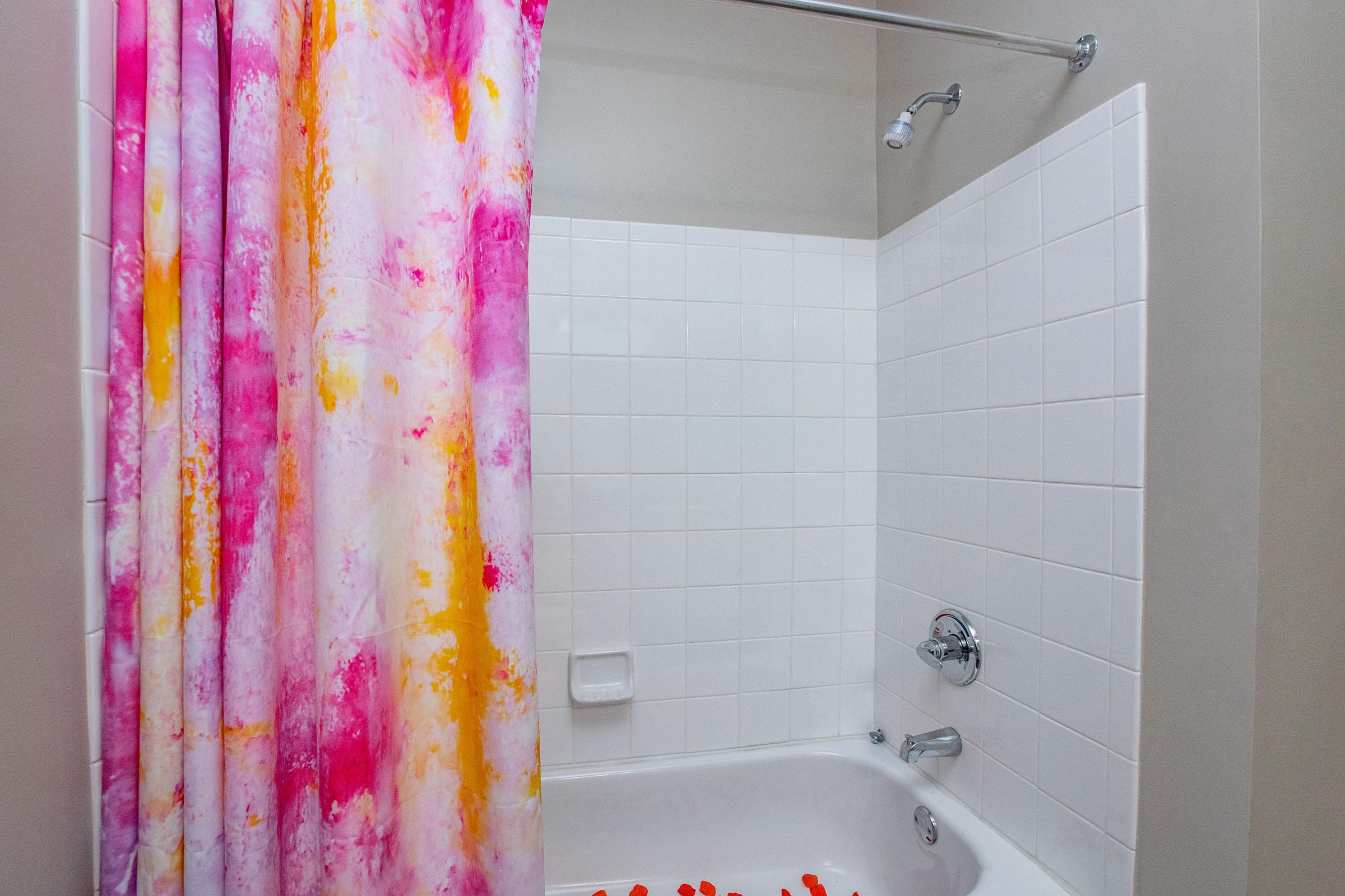 A bathroom with a pink and orange patterned shower curtain. The shower area features white tiled walls and a chrome showerhead. There is a white bathtub with a chrome faucet, and a soap dish built into the wall. Soft, warm lighting enhances the clean and inviting appearance of the space.