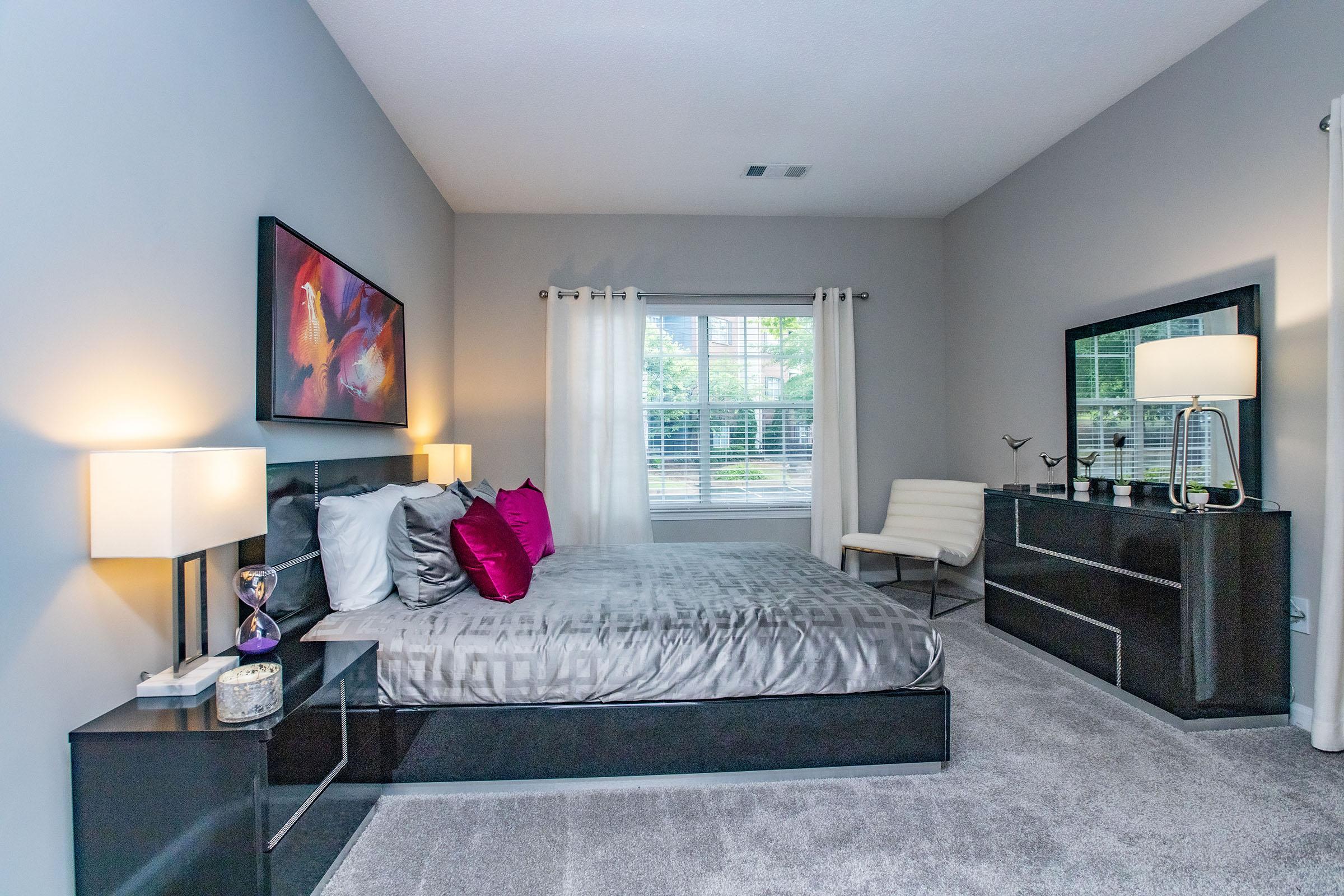 A modern bedroom featuring a gray color scheme. It has a large bed with decorative pillows in white and pink, two stylish lamps on nightstands, a sleek dresser, and a mirror. Natural light comes through the window, and the floor is covered in soft gray carpet. Wall art adds a pop of color to the space.