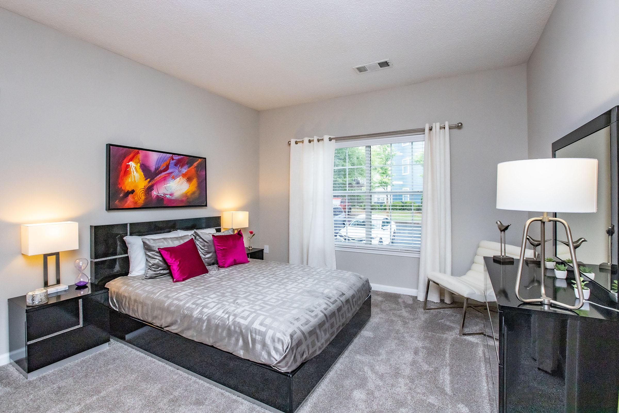 Modern bedroom featuring a large bed with decorative pillows, a colorful abstract painting above the bed, and stylish bedside lamps. A window with sheer curtains lets in natural light, and a black dresser and chair complete the contemporary decor. The carpeted floor adds warmth to the space.