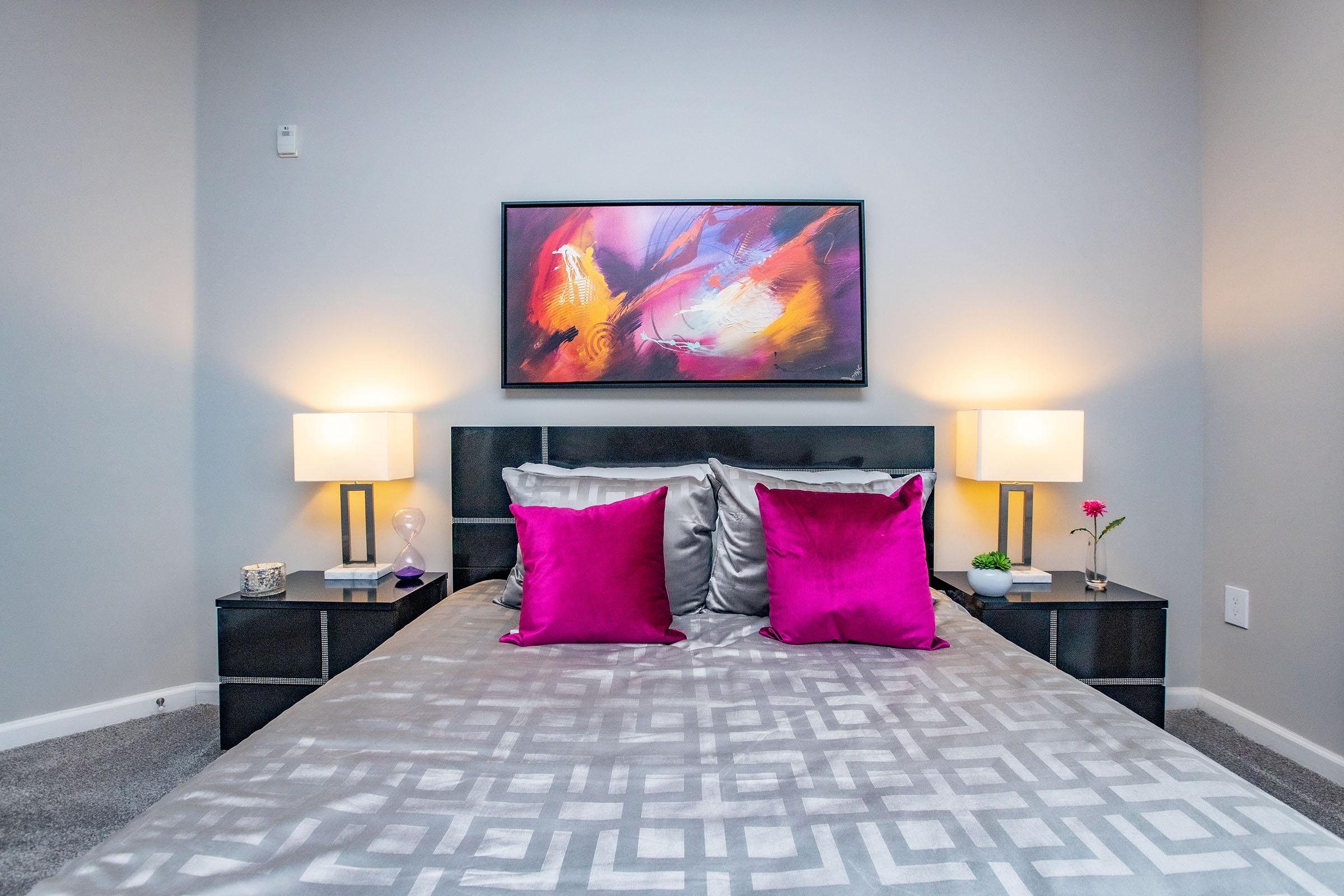 A modern bedroom featuring a bed with a silver duvet and vibrant pink pillows. On either side of the bed are nightstands with lamps. Above the bed hangs an abstract painting with bold colors. The walls are a neutral gray, and a small plant and flower are placed decoratively on the nightstands.