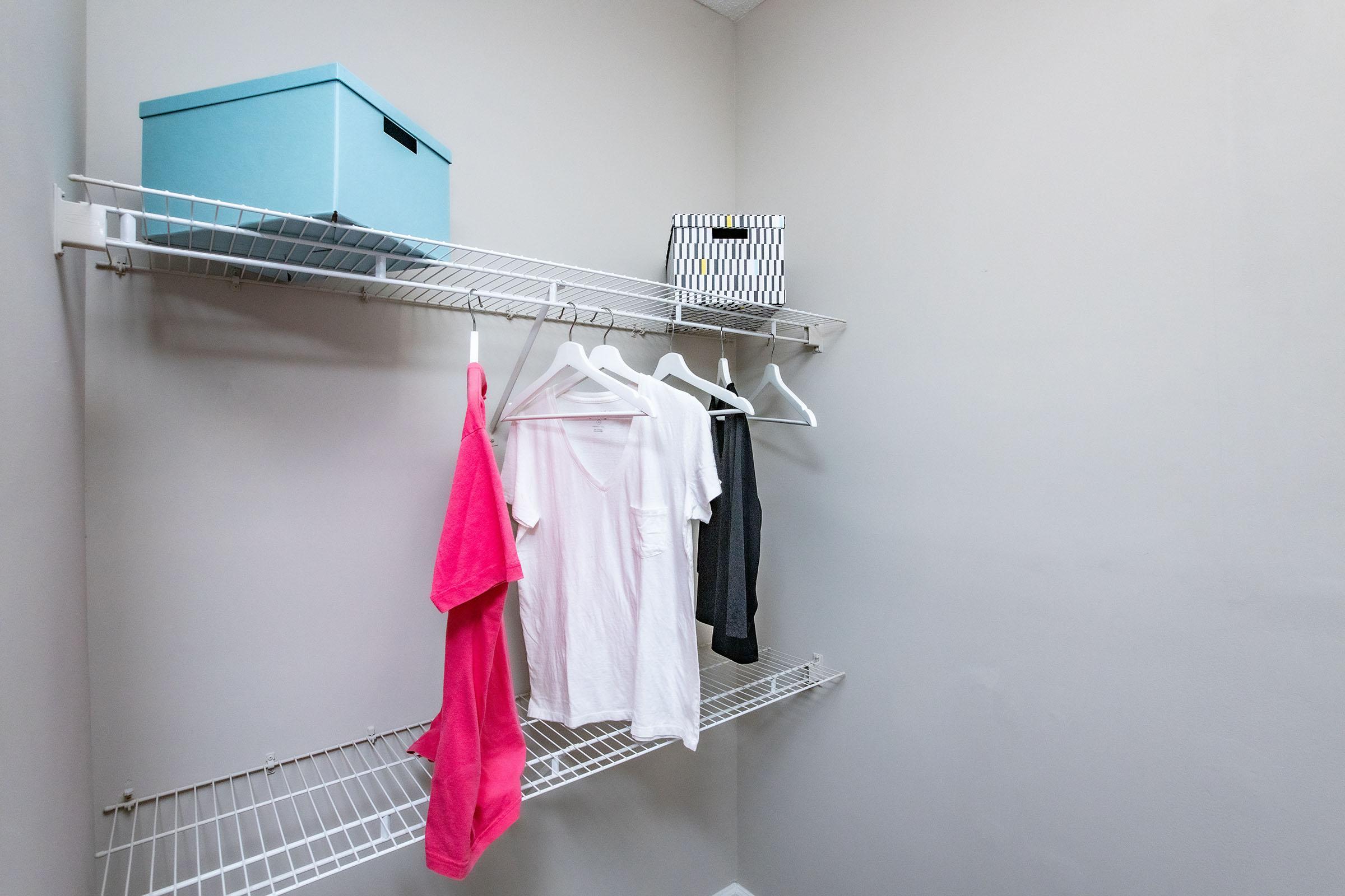 A minimalistic laundry room with two wall-mounted wire shelves. The top shelf holds a light blue storage box and a black-and-white striped box. The bottom shelf features three white hangers with clothing: a pink towel, a white shirt, and a dark gray shirt. The walls are painted a light gray.