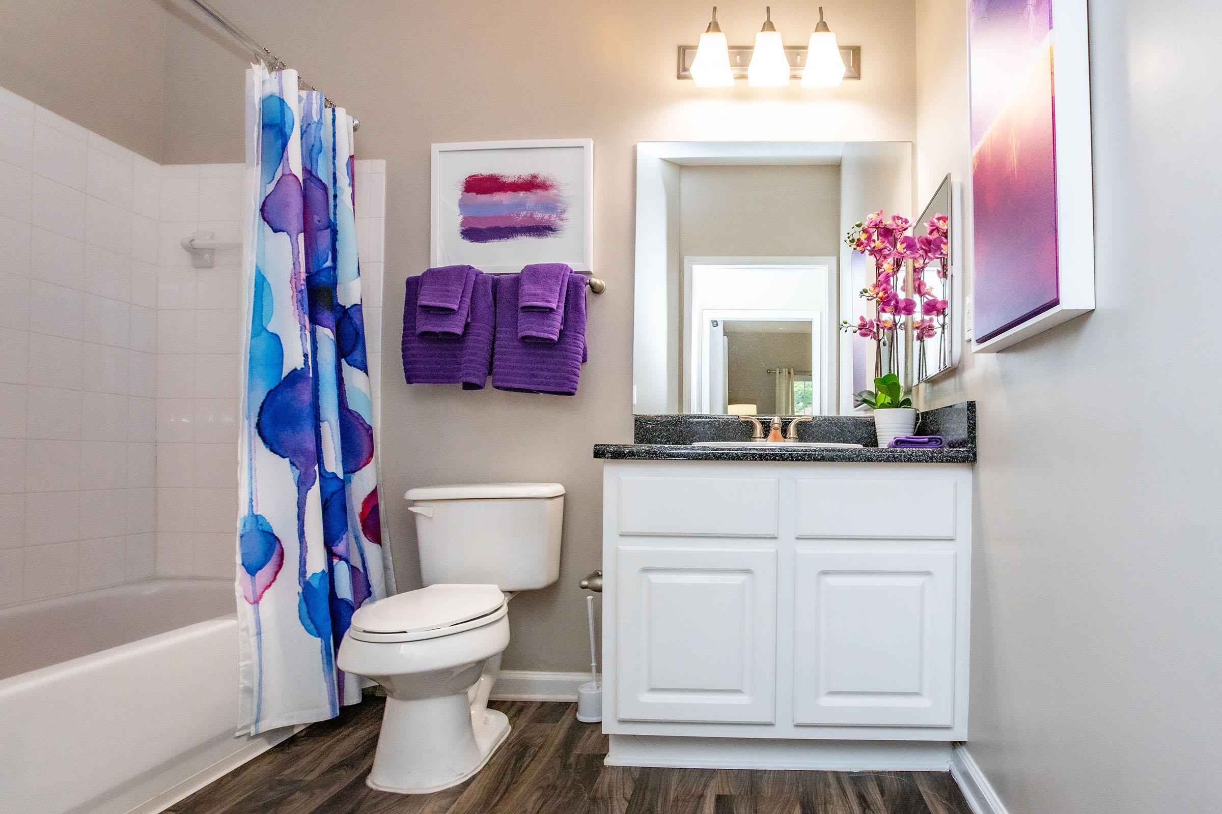 A modern bathroom featuring a white toilet, a dark countertop with a mirror above, and a colorful shower curtain with blue and purple patterns. Two purple towels hang on the wall, and a framed artwork with a gradient purple design is displayed. The floor has a wood-like appearance.