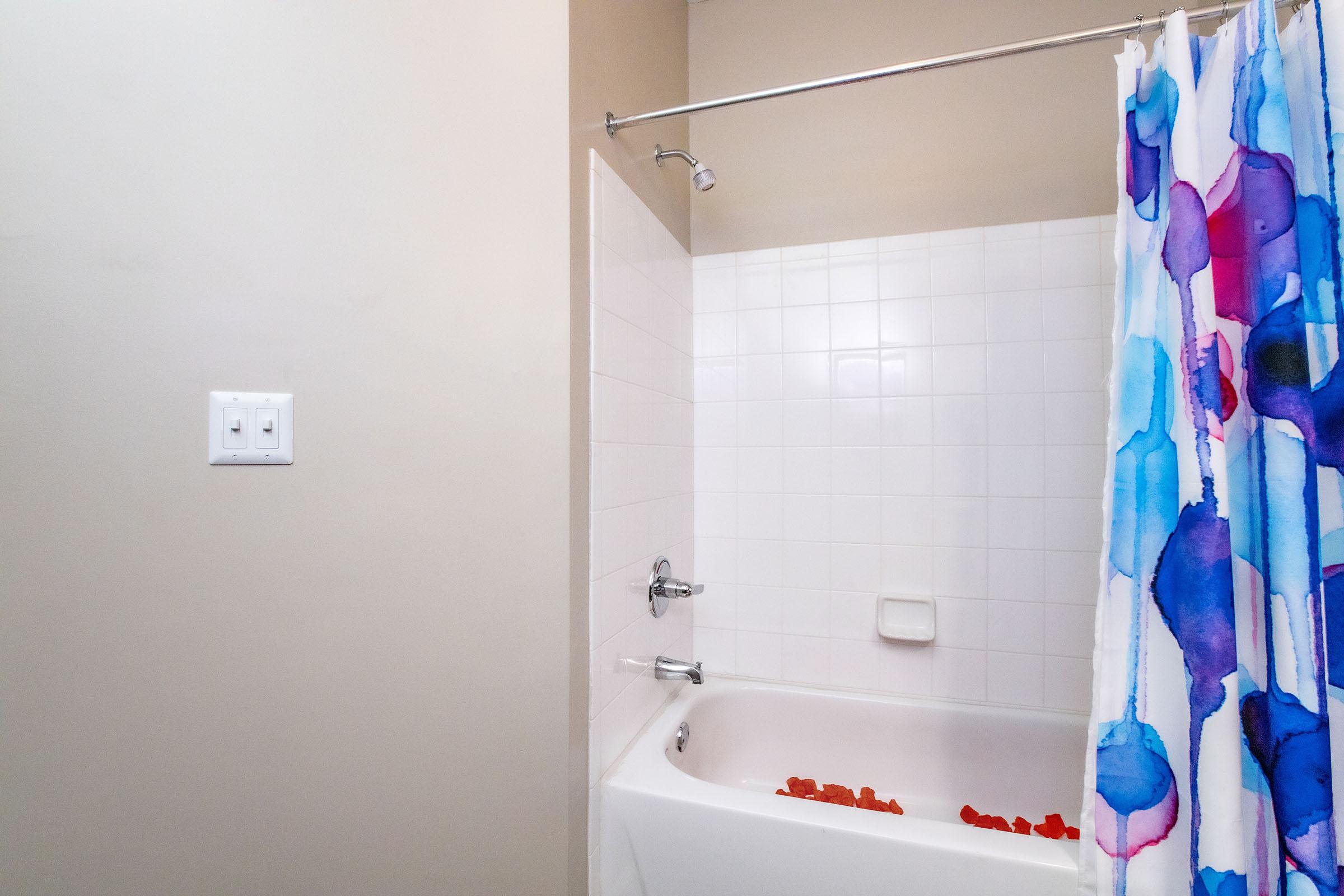 A bathroom with a bathtub partially covered by a colorful shower curtain featuring blue and purple patterns. The bathtub contains rose petals, and the wall has white tiles. A light switch is visible on the left side of the image. The overall space has a simple and clean aesthetic.