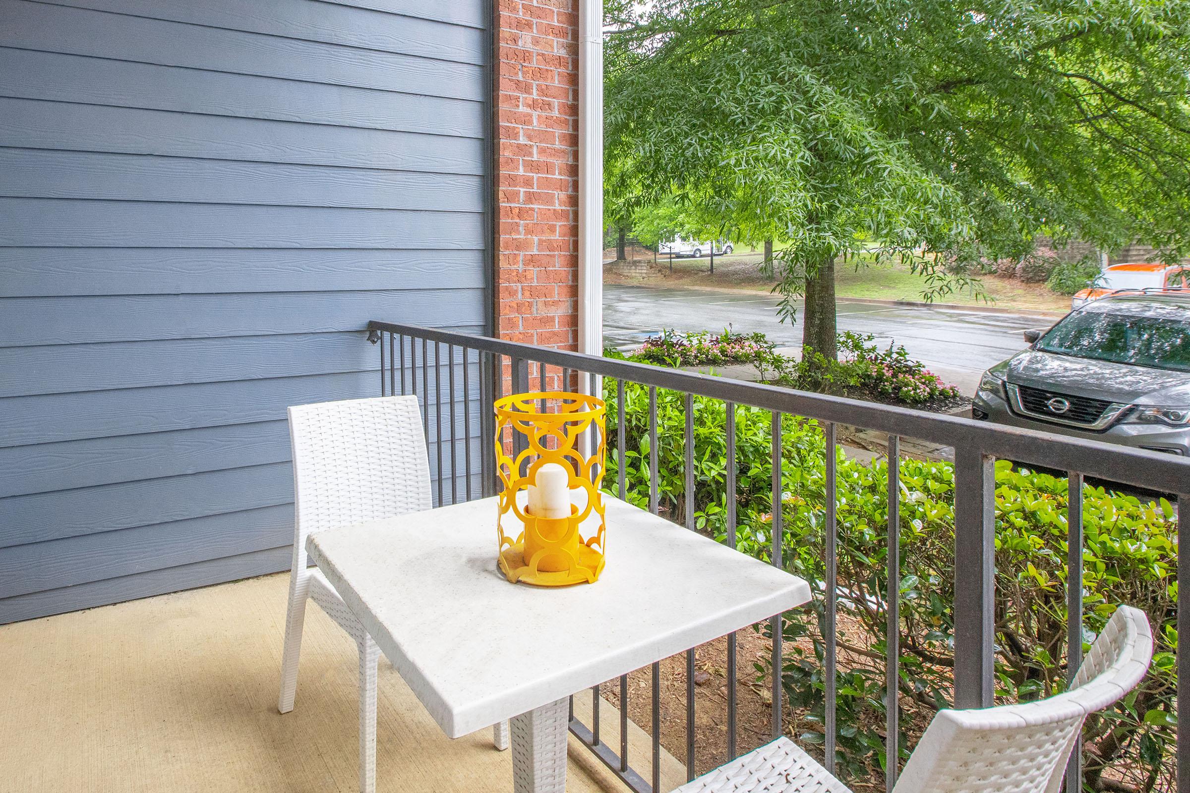 A small outdoor balcony features a white table and two white chairs. On the table, there is a yellow decorative lantern with a candle. Surrounding the balcony are green shrubs and trees, with a glimpse of a parked car and a road in the background. The wall is a soft blue, contrasting with the brick.