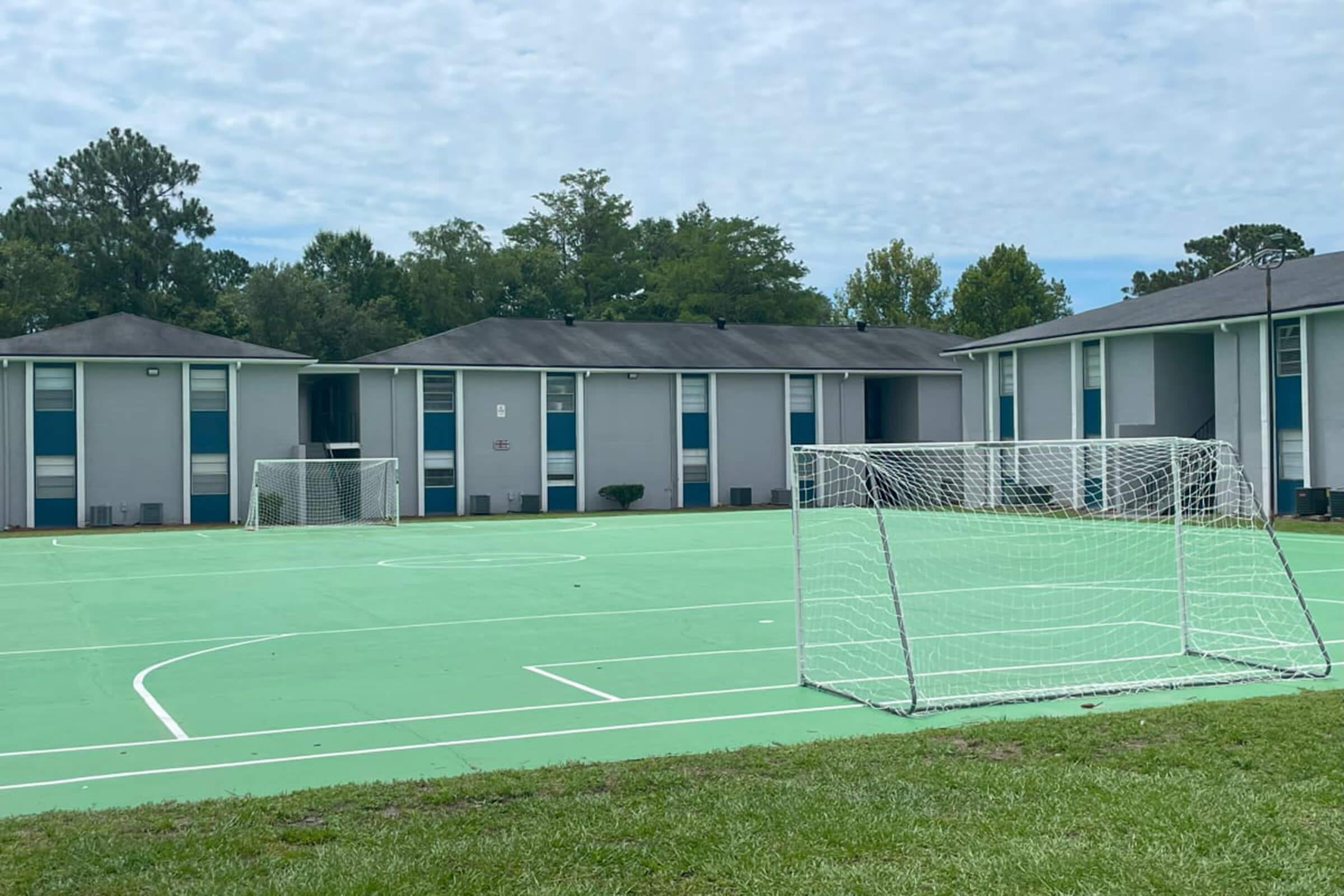A green playground with soccer goals at either end, surrounded by apartment buildings. The sky is cloudy, and there are trees in the background. The surface is marked for both soccer and other sports activities.
