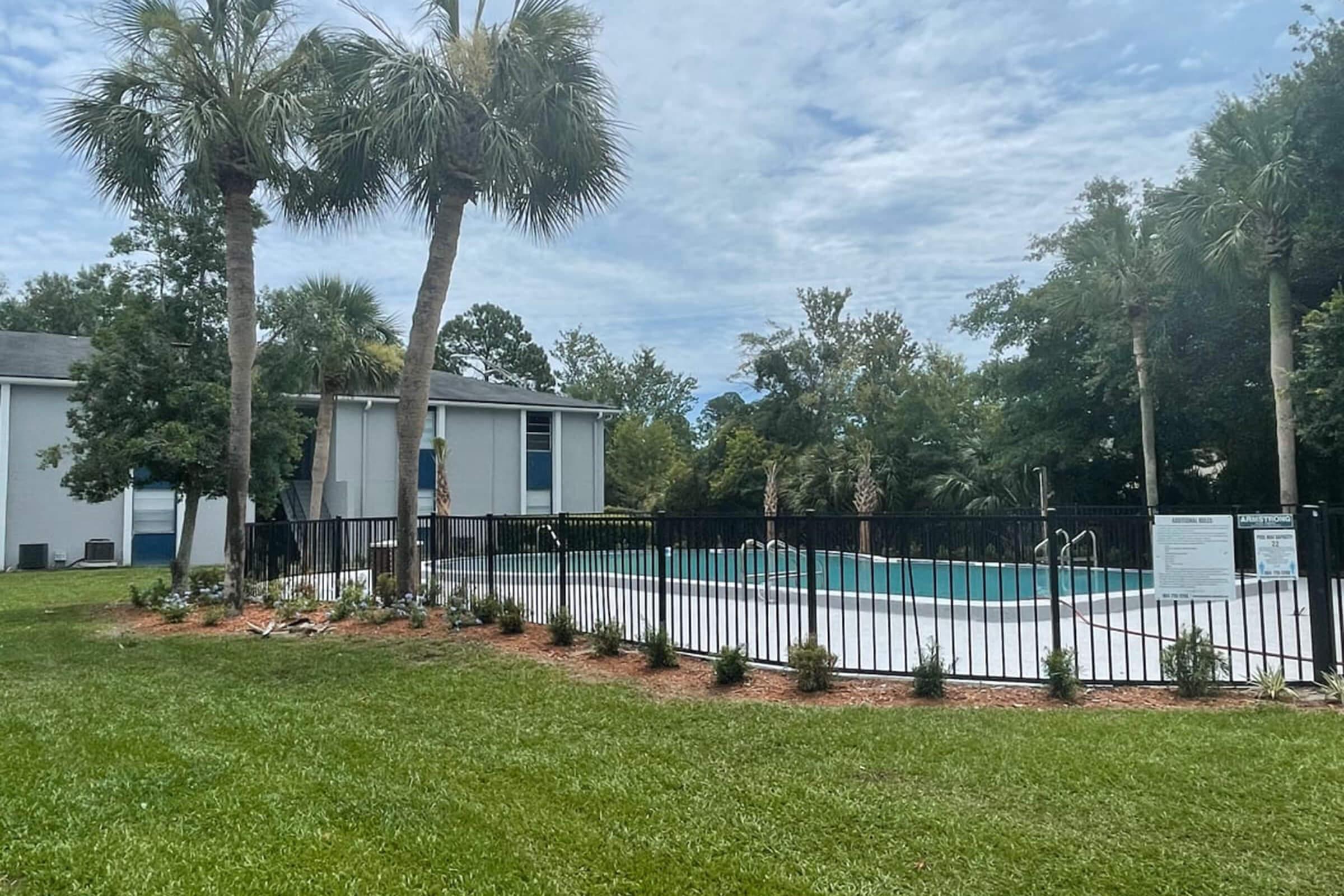 A peaceful scene featuring a fenced swimming pool surrounded by lush green grass and palm trees. In the background, a building is visible, partially obscured by trees. The sky is overcast, adding a calm ambiance to the setting.