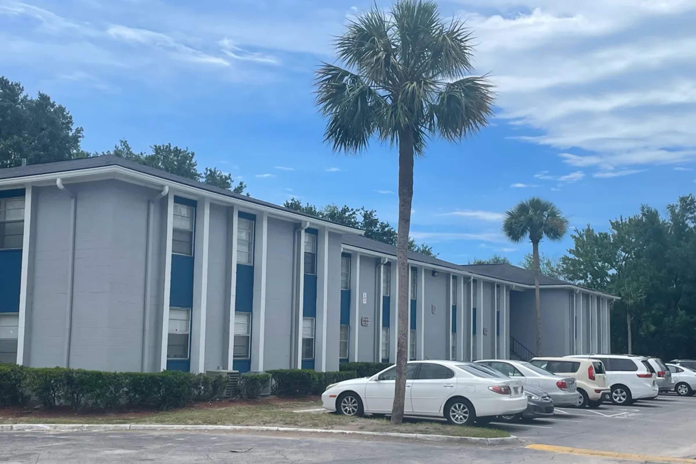 A two-story gray apartment building with blue accents, surrounded by palm trees. Several cars are parked in a lot in front of the building. The sky is partly cloudy, suggesting a sunny day.
