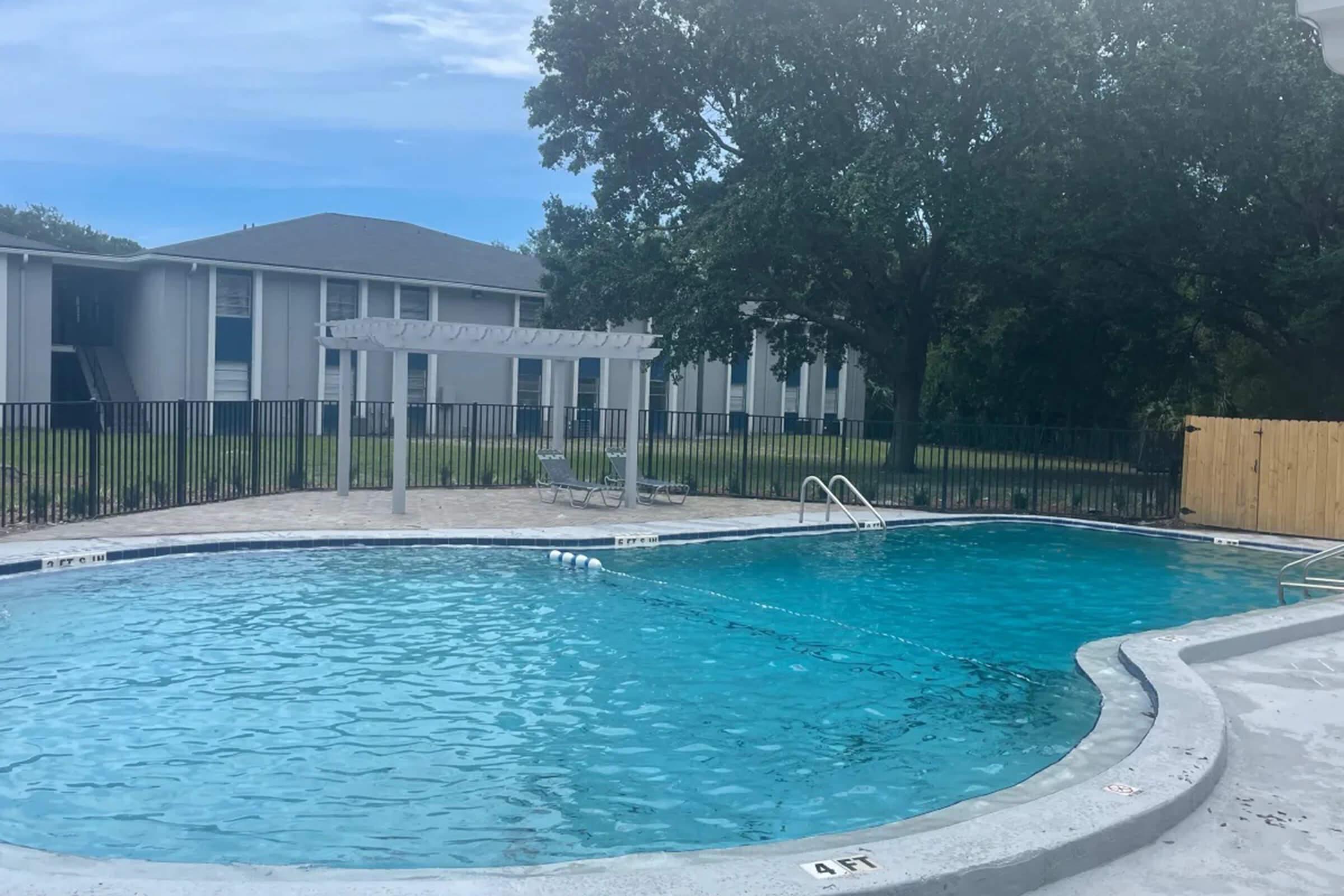 A serene swimming pool area featuring two pools with clear blue water, surrounded by a fence and shaded by trees. In the background, there are buildings visible, and some lounge chairs are placed nearby. The sky is mostly clear with a few clouds.
