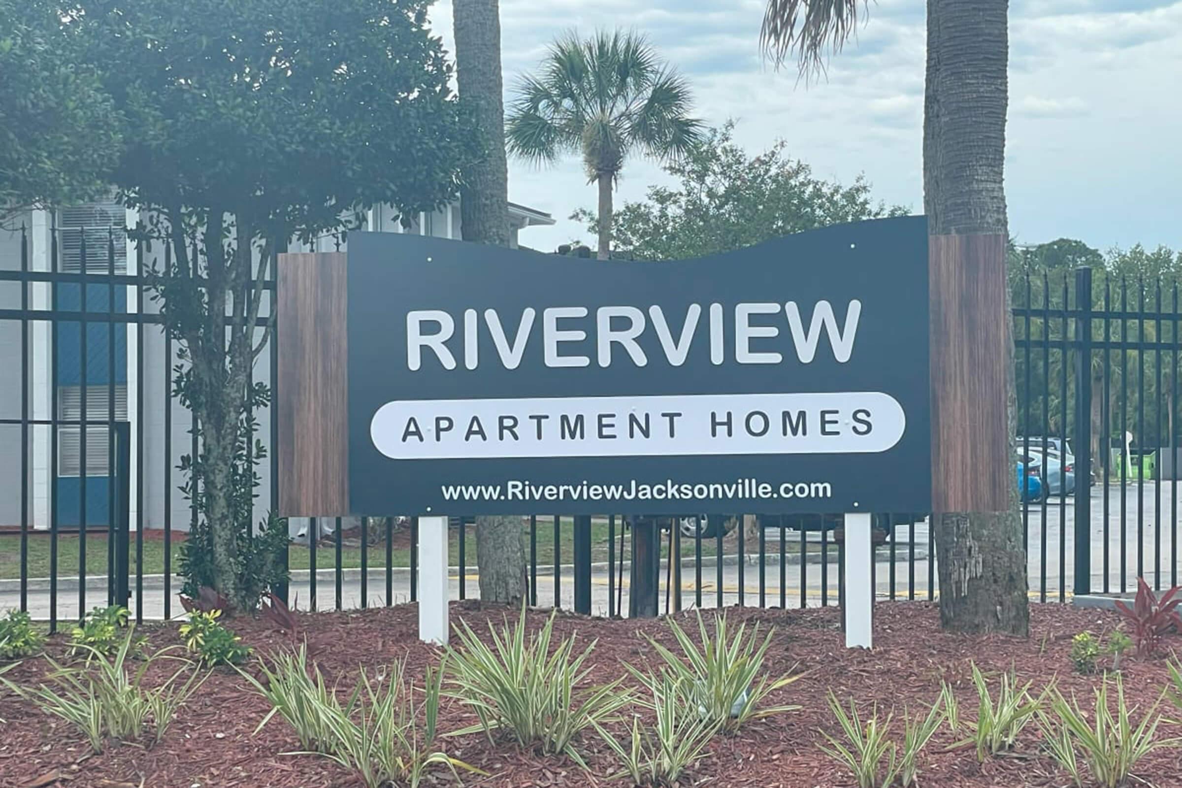 Sign for Riverview Apartment Homes, featuring a modern design with the text "RIVERVIEW" prominently displayed. The sign is surrounded by landscaped areas with plants and palm trees, and a fence visible in the background. The website address "www.RiverviewJacksonville.com" is listed at the bottom.
