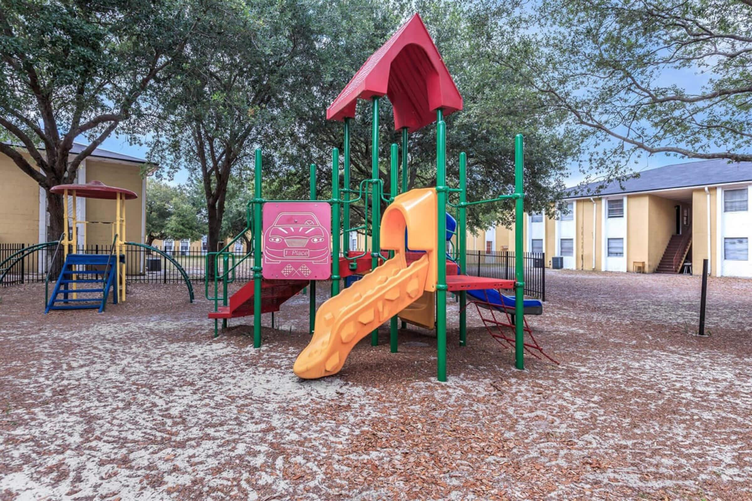 A colorful playground featuring a climbing structure with a slide, swings, and a playhouse, situated in a sandy area shaded by trees. Two residential buildings are visible in the background, creating a welcoming environment for children to play.