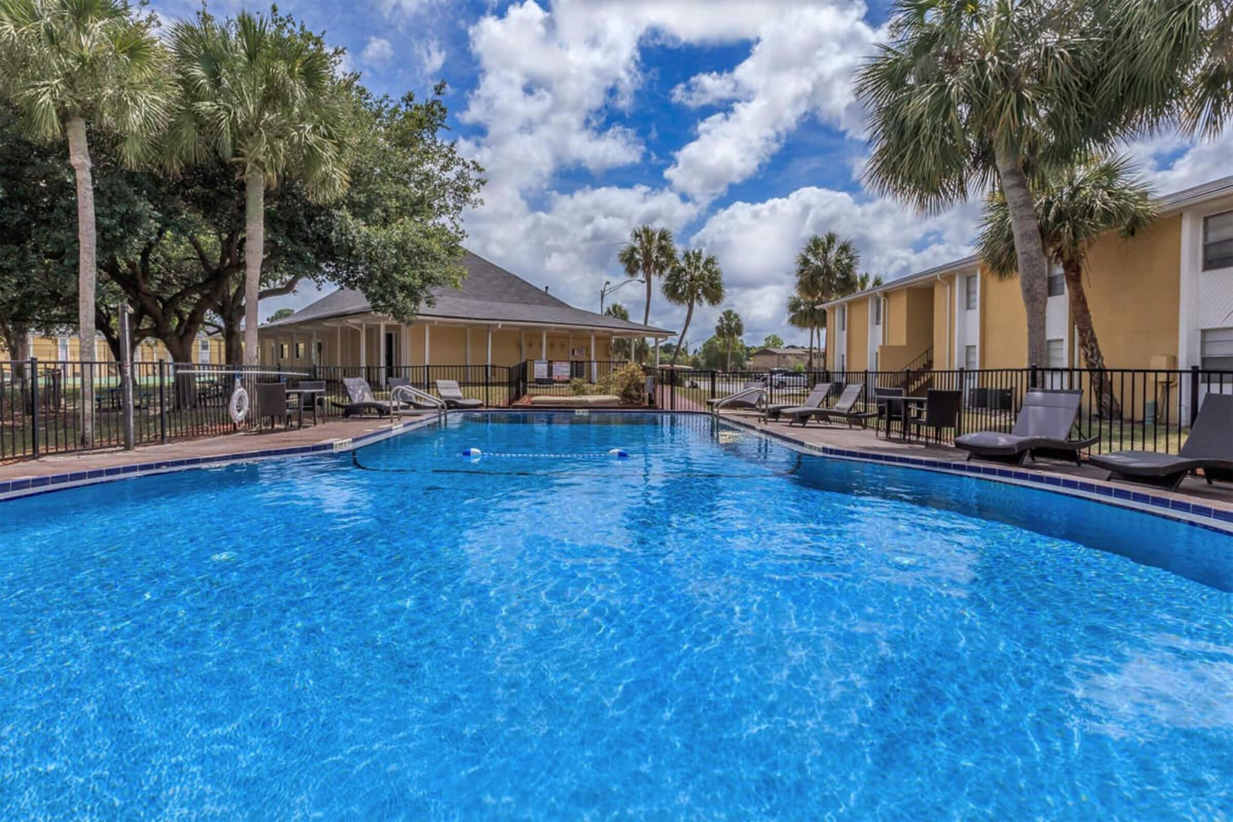 A sparkling blue swimming pool surrounded by palm trees and lounge chairs, with a residential building in the background under a cloudy sky. The scene conveys a relaxing outdoor atmosphere, ideal for leisure and enjoyment.