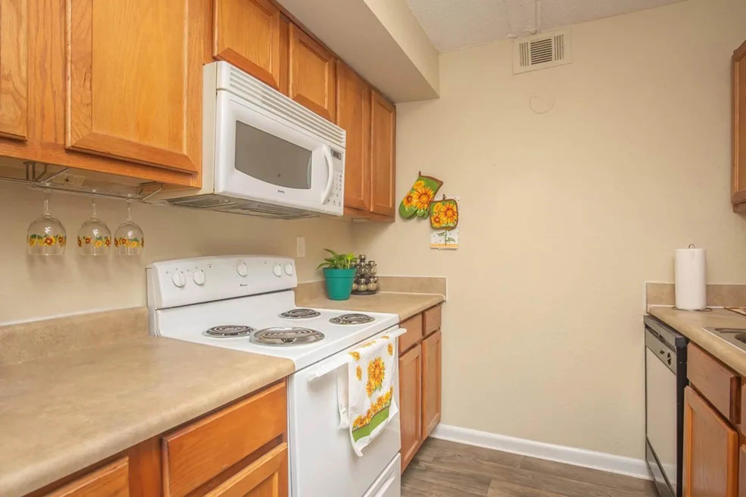 A cozy kitchen featuring wooden cabinets, a white microwave, and a white stove with four burners. The countertops are beige, and there are hanging glasses above the sink. A small potted plant and various kitchen items are visible, along with a decorative towel hanging from the stove. The walls are a light beige color.