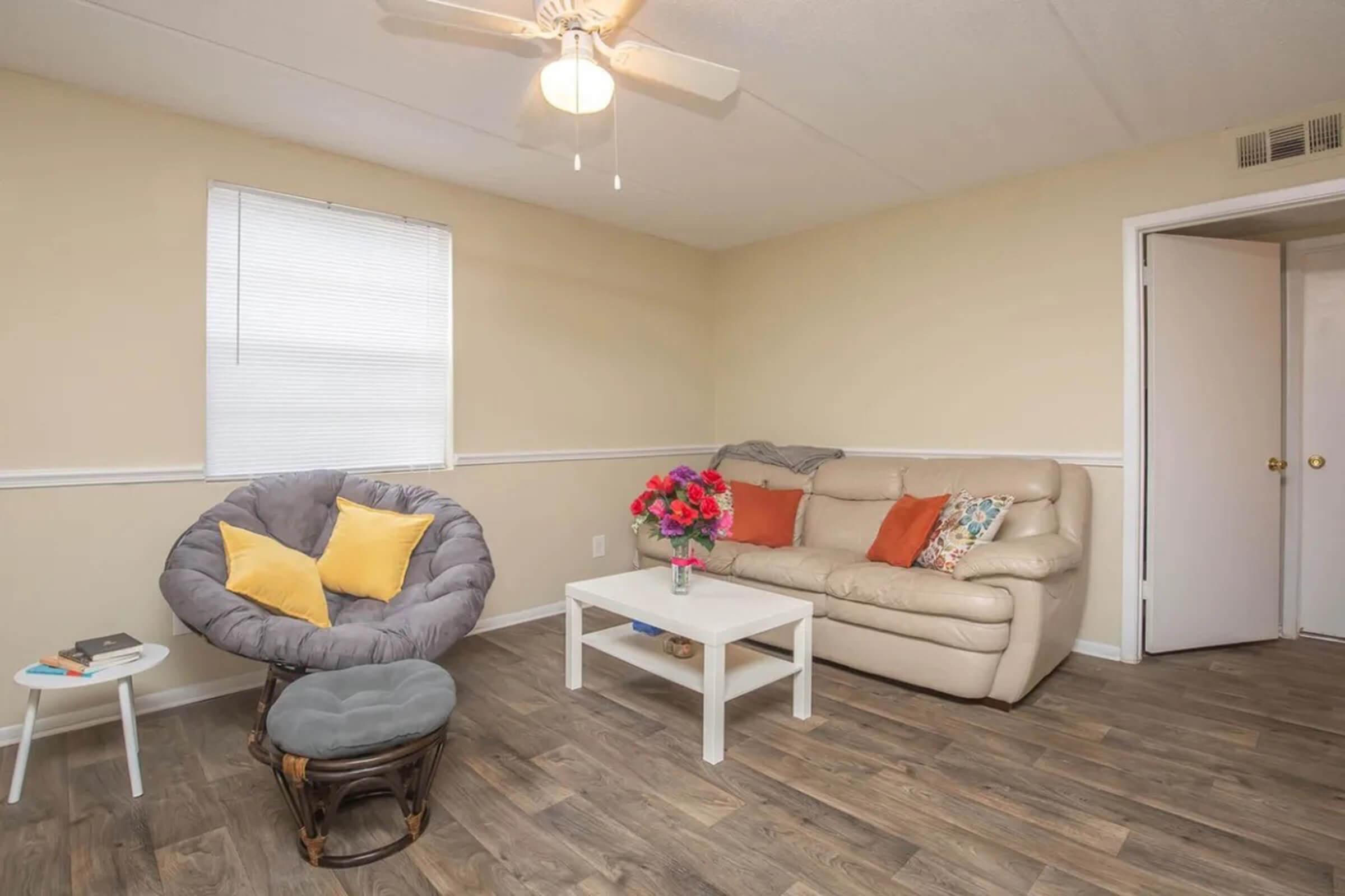 Living room with beige walls and a ceiling fan. A light-colored couch with decorative pillows is positioned against the wall. A small white coffee table sits in front of the couch, with a vase of flowers on top. A cozy, circular chair with bright cushions is nearby, and there's a door leading to another room.