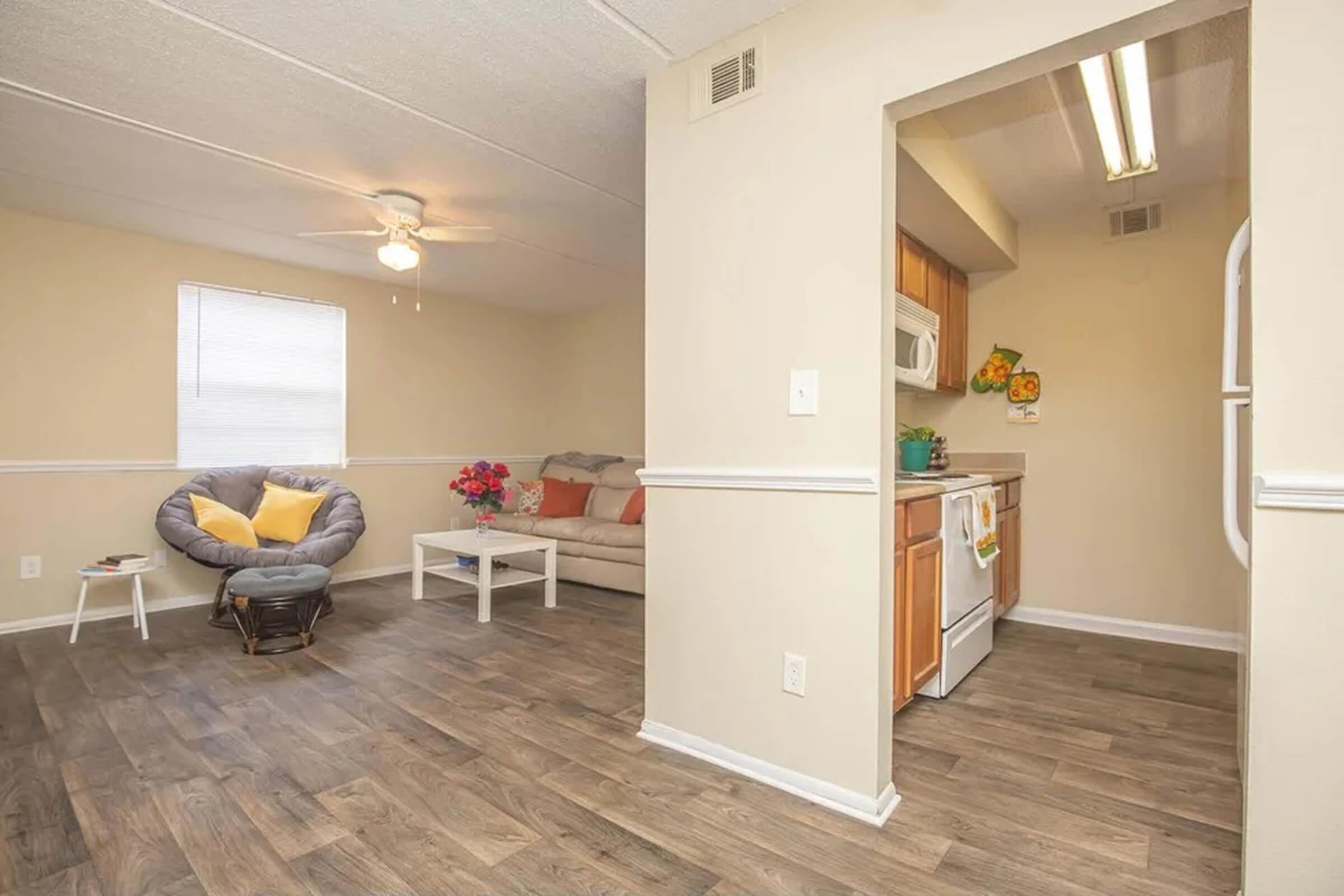 A cozy living room leading to a kitchen, featuring a beige couch with colorful pillows, a round chair, a coffee table with flowers, and a ceiling fan. The kitchen has white cabinetry, an oven, and a well-organized countertop, with light-colored walls and wood-look flooring throughout. Natural light fills the space.
