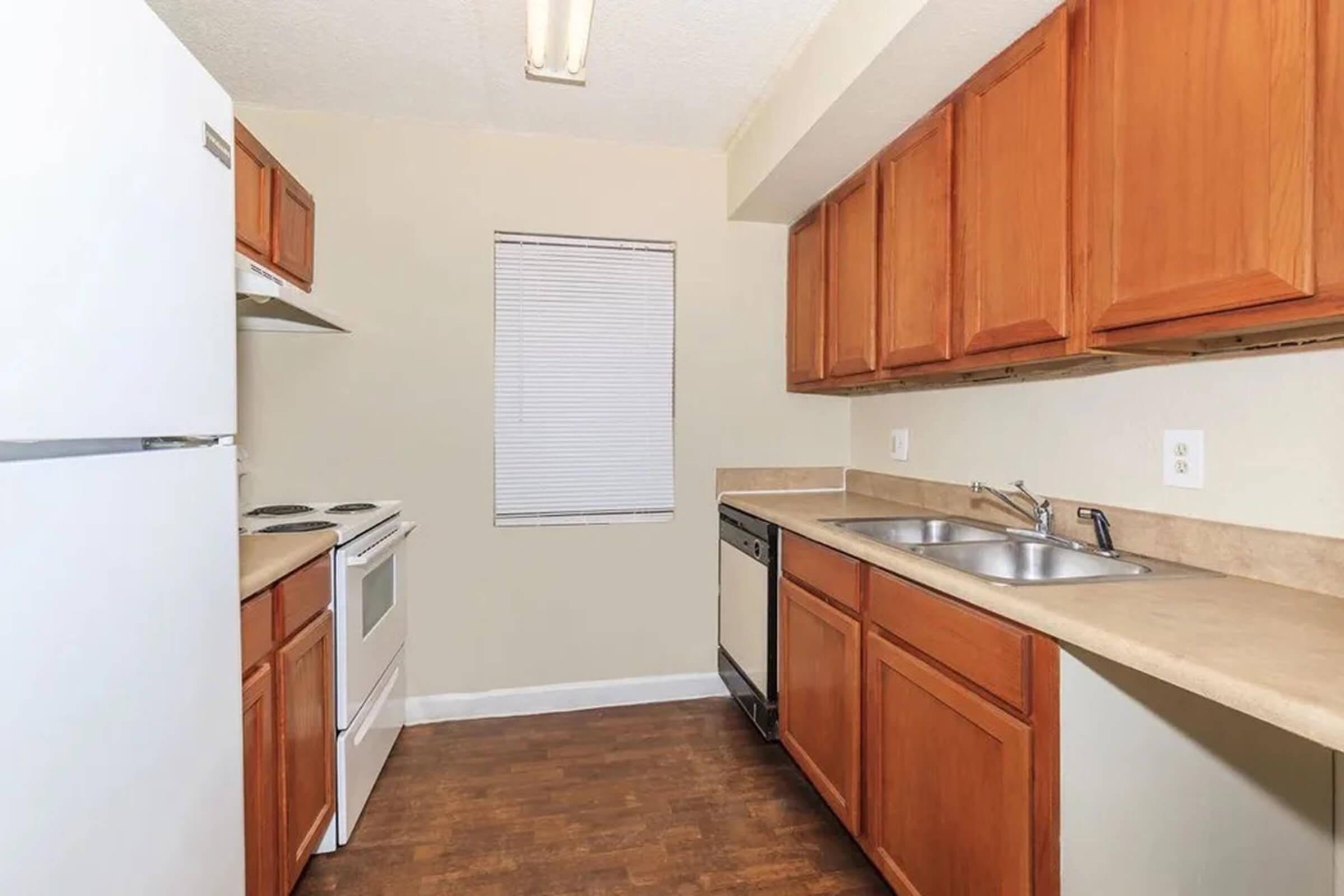 A modern kitchen featuring wooden cabinets, a countertop, and standard appliances including a refrigerator, oven, and dishwasher. There's a double sink and a window with blinds, providing natural light. The flooring is a hardwood-like material.