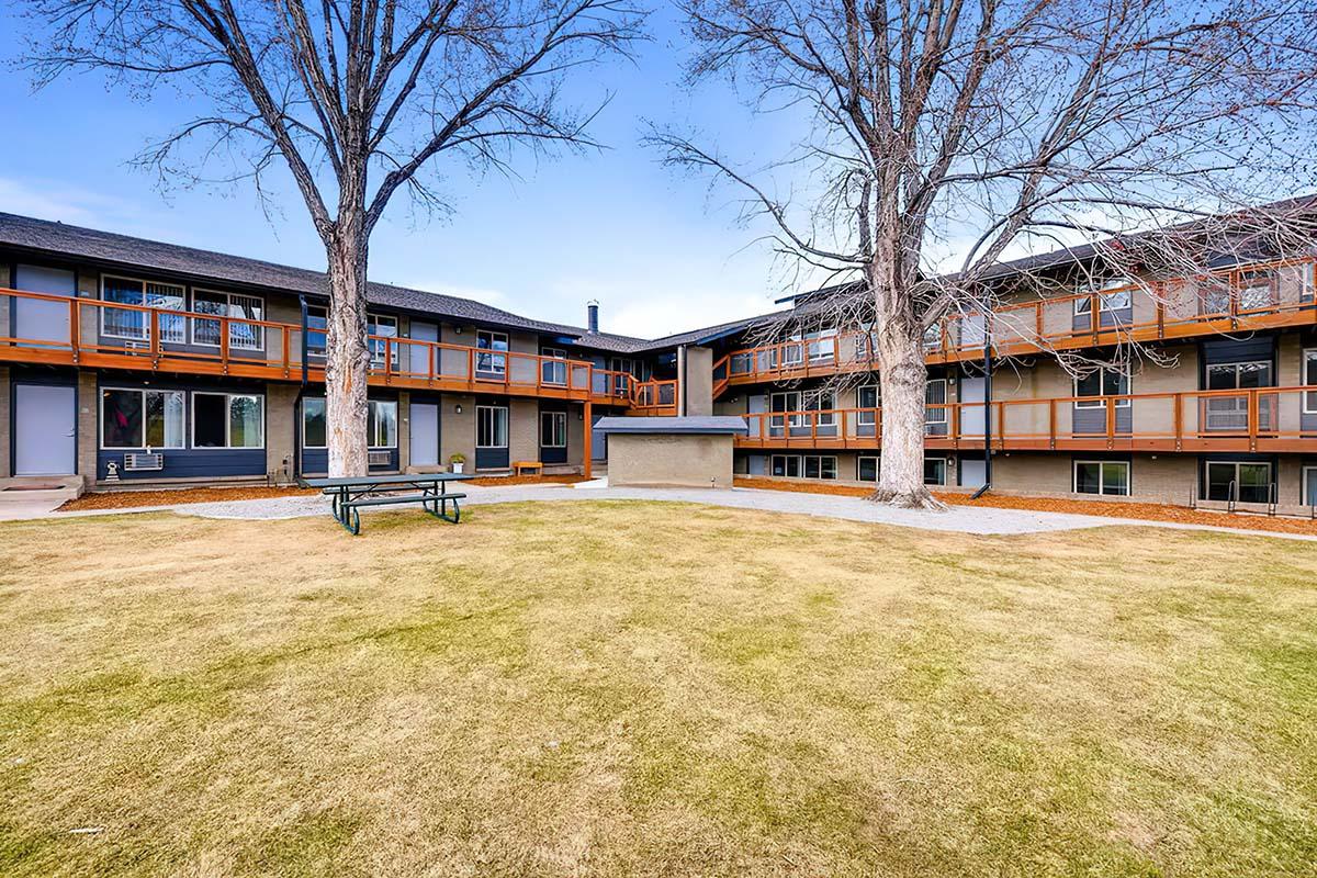 A view of an outdoor area between two apartment buildings featuring grassy space, a picnic table, and two large trees