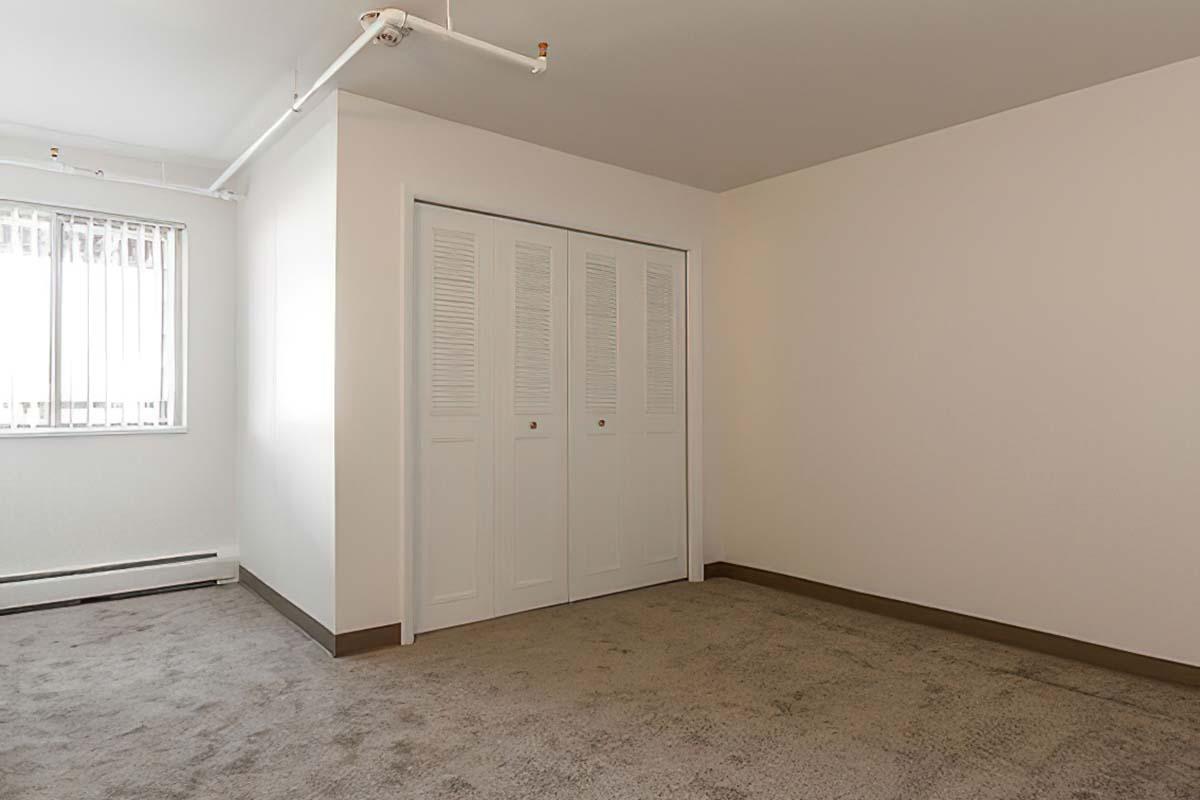 A vacant room with beige walls and a light brown carpet. There is a large closed sliding door closet on one wall and a window with vertical blinds allowing natural light to enter.