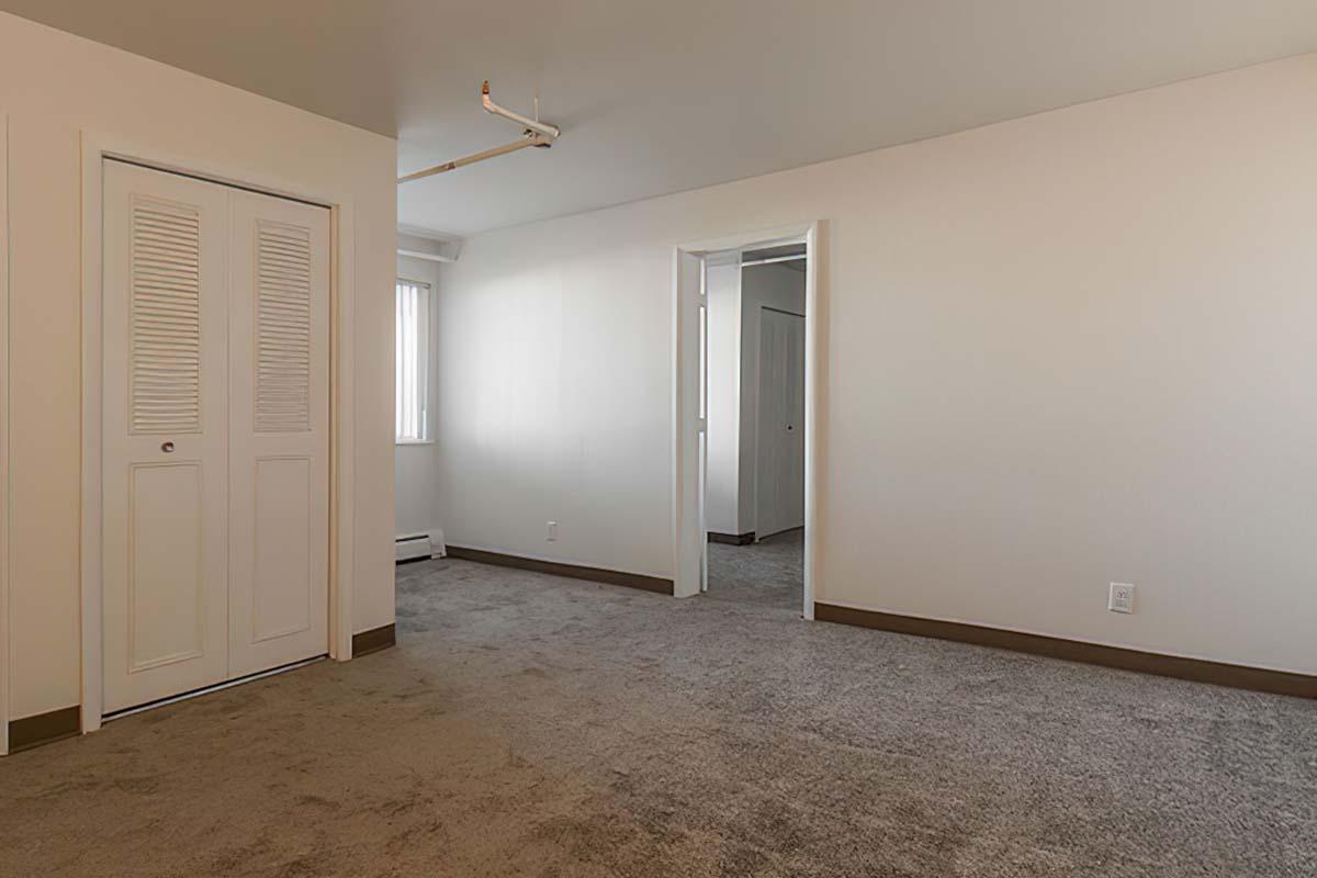 A spacious, empty room featuring light-colored walls and carpeted flooring. The room includes a closet with double doors on the left and a doorway leading to another area.