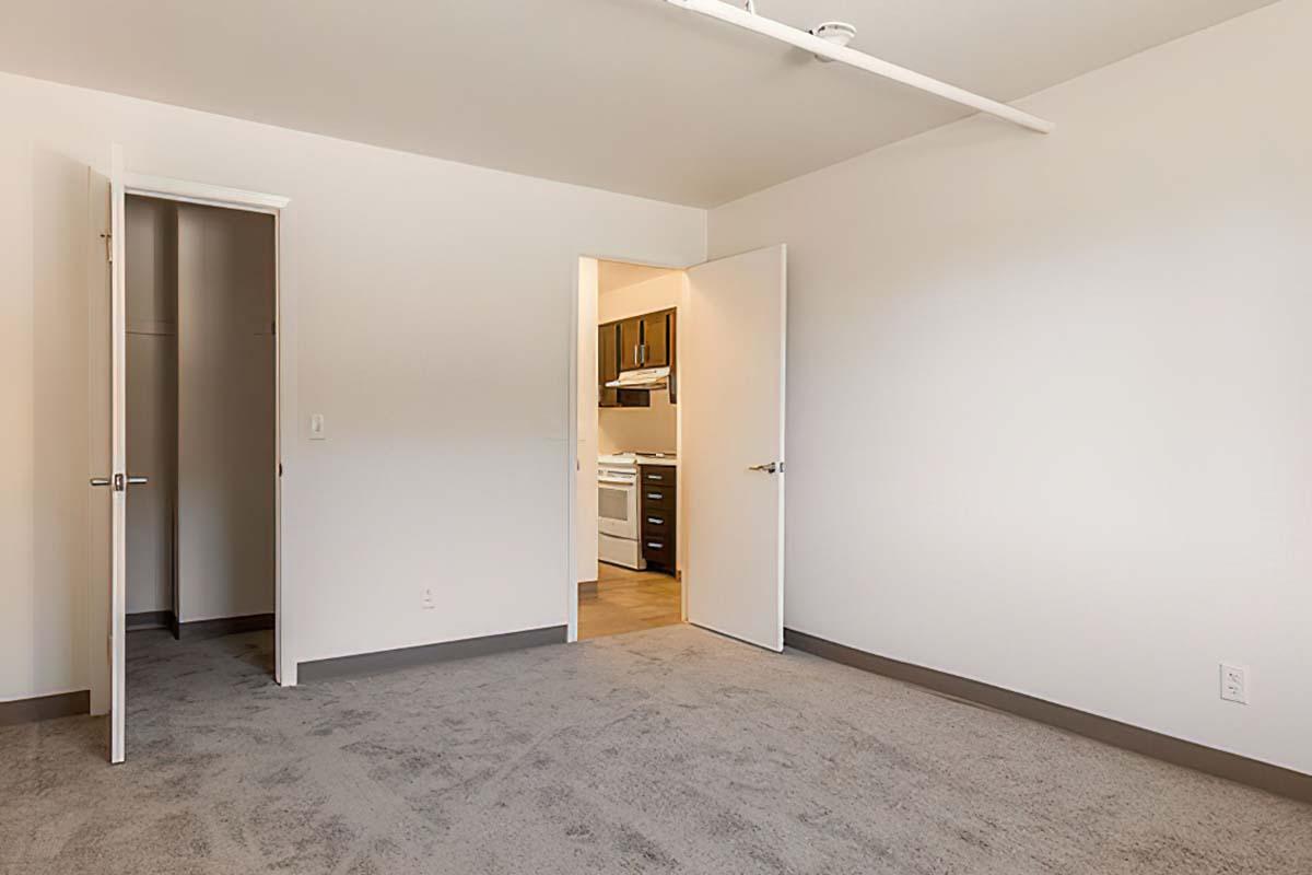 A spacious, empty room with light gray carpet, featuring two open doors. One door leads to a closet, while the other opens to a kitchen area with a white stove visible in the background.