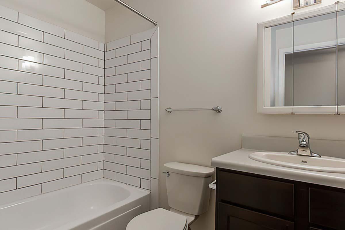 A modern bathroom featuring a white subway tile shower wall, a bathtub, a white toilet, and a dark wood vanity with a sink.