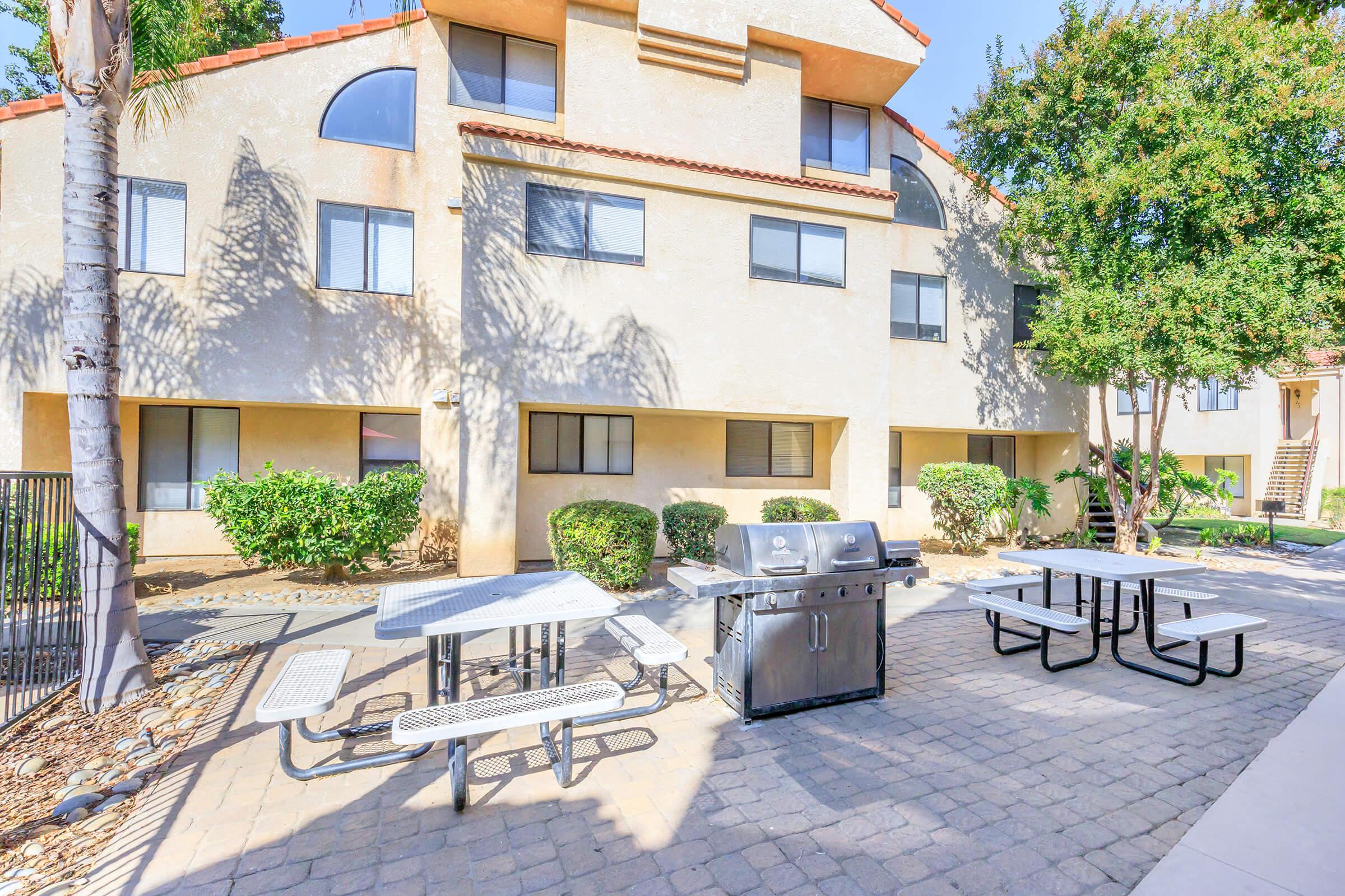 A landscaped outdoor area featuring a grill, picnic tables, and surrounding shrubs, situated next to a multi-story building with large windows and a bright sky.