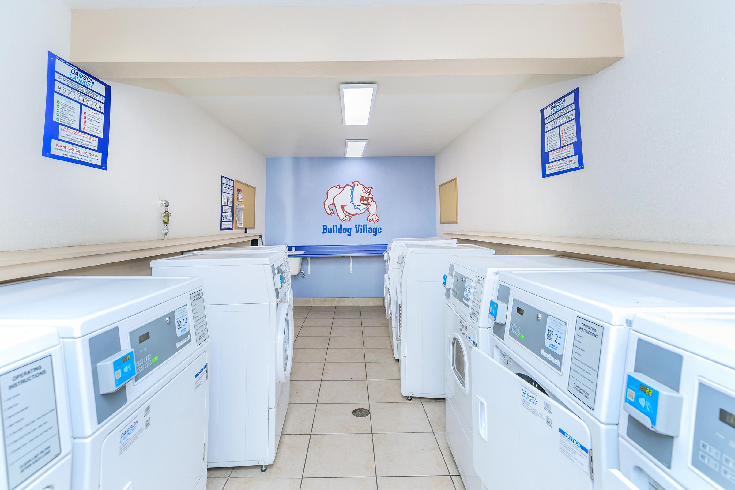 Interior view of a laundry room named "Bulldog Village," featuring several white washing machines and dryers. Brightly lit with smooth tiled flooring and beige walls. Informational signs are posted on the walls, providing details about laundry usage. A workspace is visible at the end of the room.