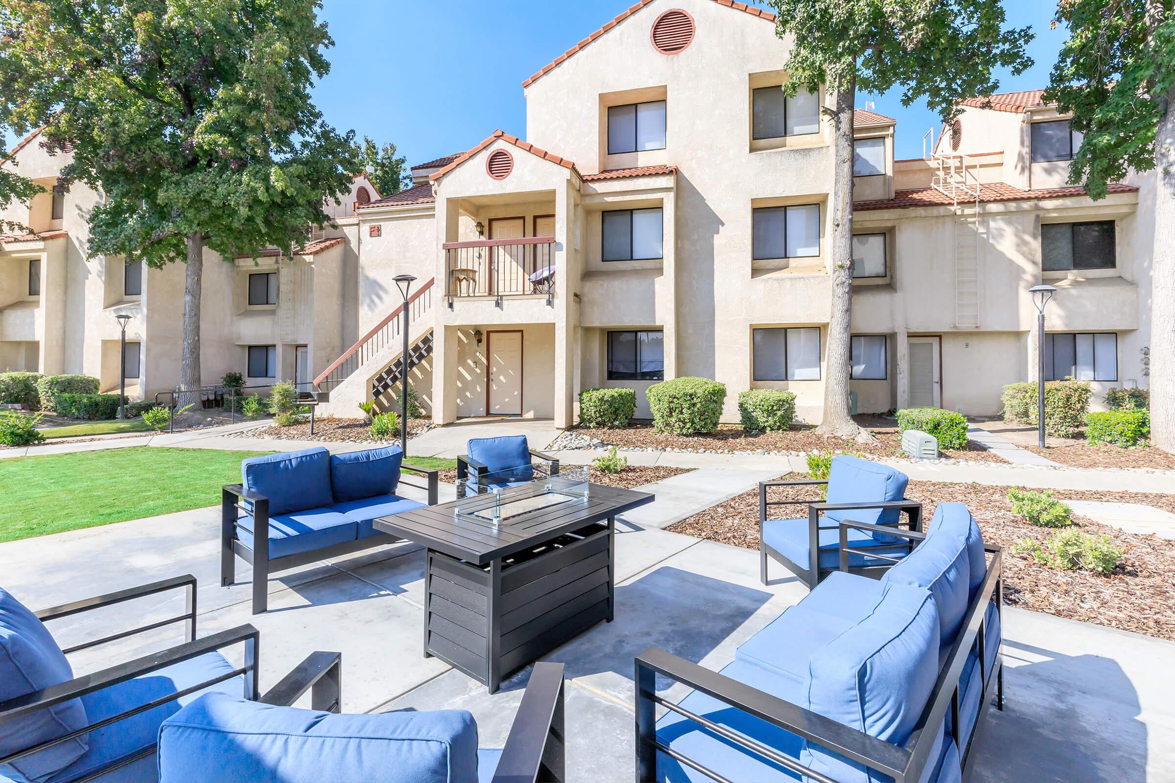 A landscaped courtyard featuring a modern outdoor seating area with blue chairs and a black table. Surrounding the seating are well-maintained shrubs and trees, alongside beige apartment buildings with balconies. The sky is clear and sunny, creating a welcoming atmosphere.