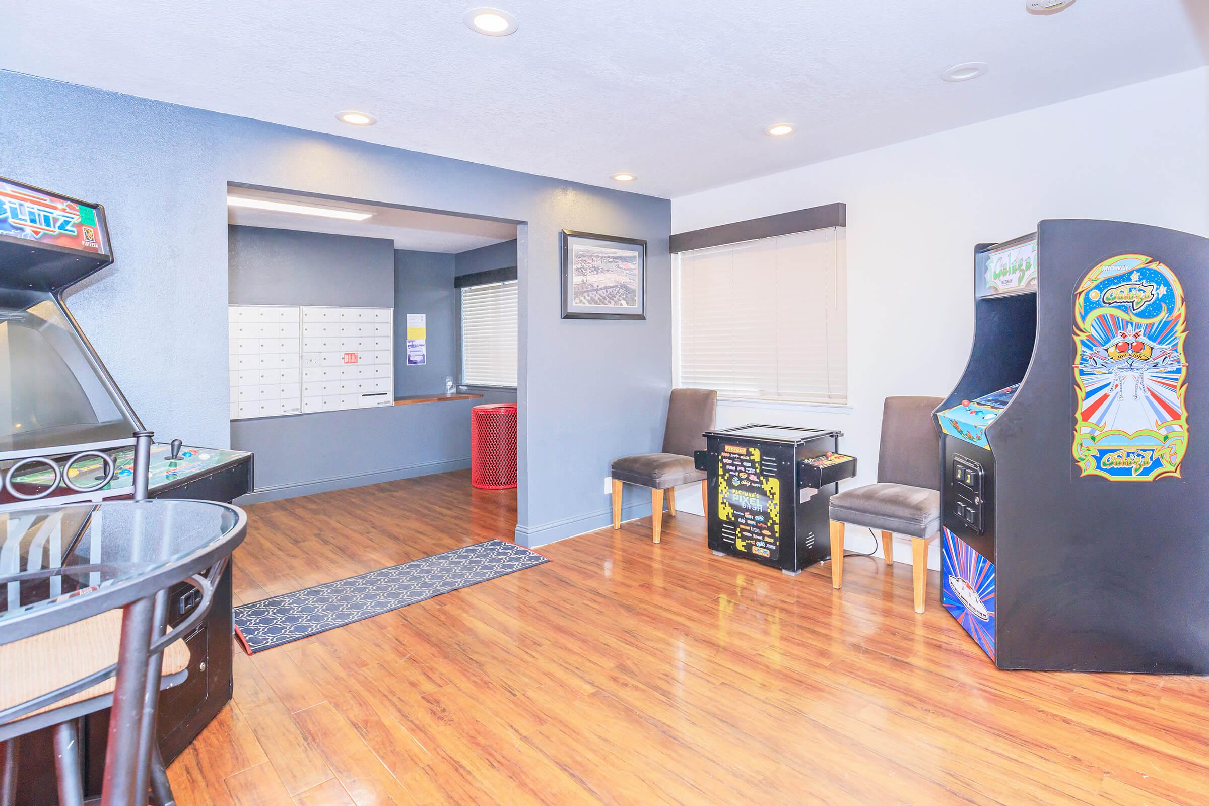 A cozy game room featuring vintage arcade machines, a small table game, and seating. The wood flooring complements the blue walls, while a mail area with lockers is visible in the background. Natural light streams in through window blinds, creating an inviting atmosphere.