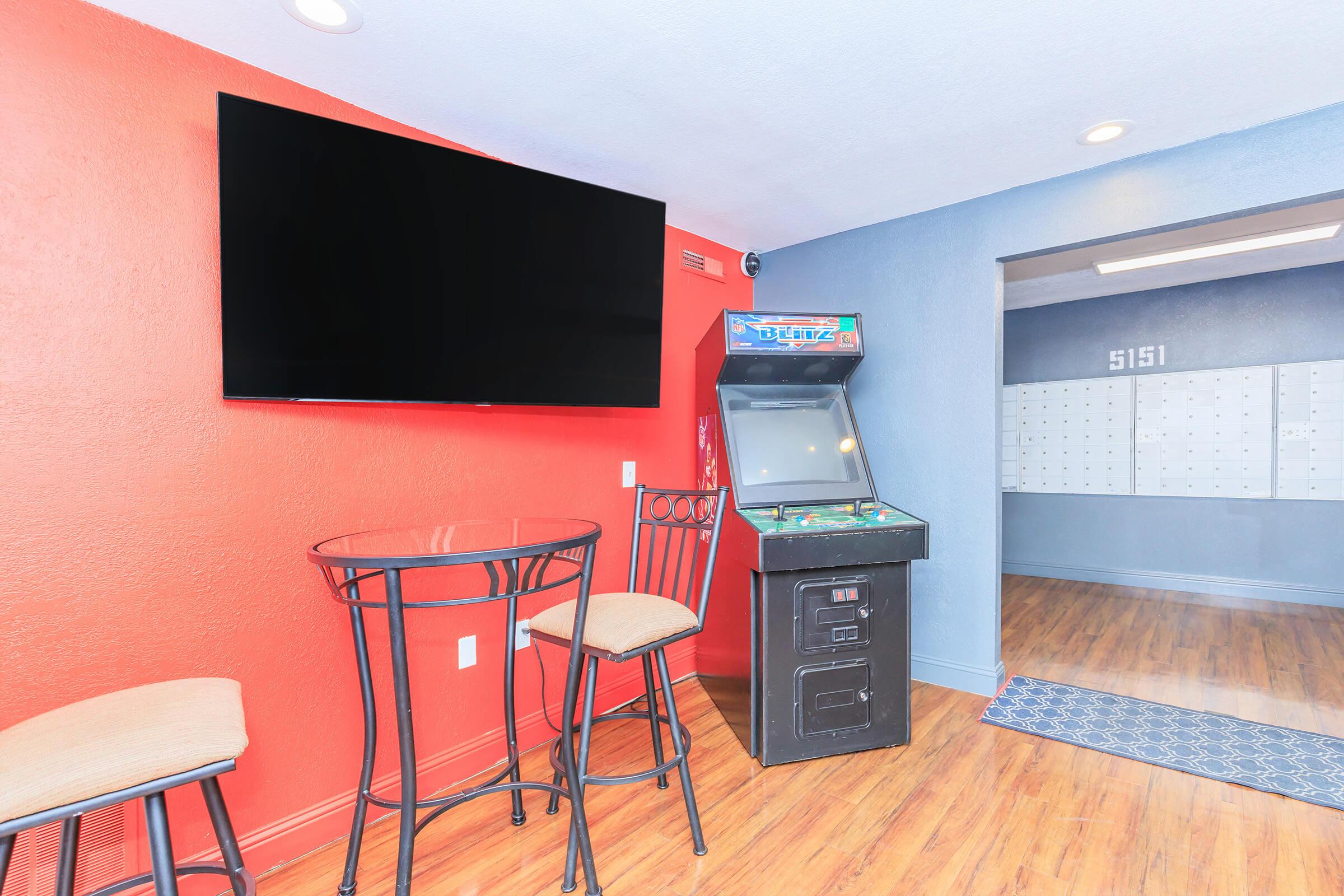A lounge area featuring a red accent wall and a blue wall. There is a flat-screen TV mounted on the wall, a round table with two bar stools, and an arcade game machine nearby. The floor is wooden, creating a cozy atmosphere for relaxation and entertainment.