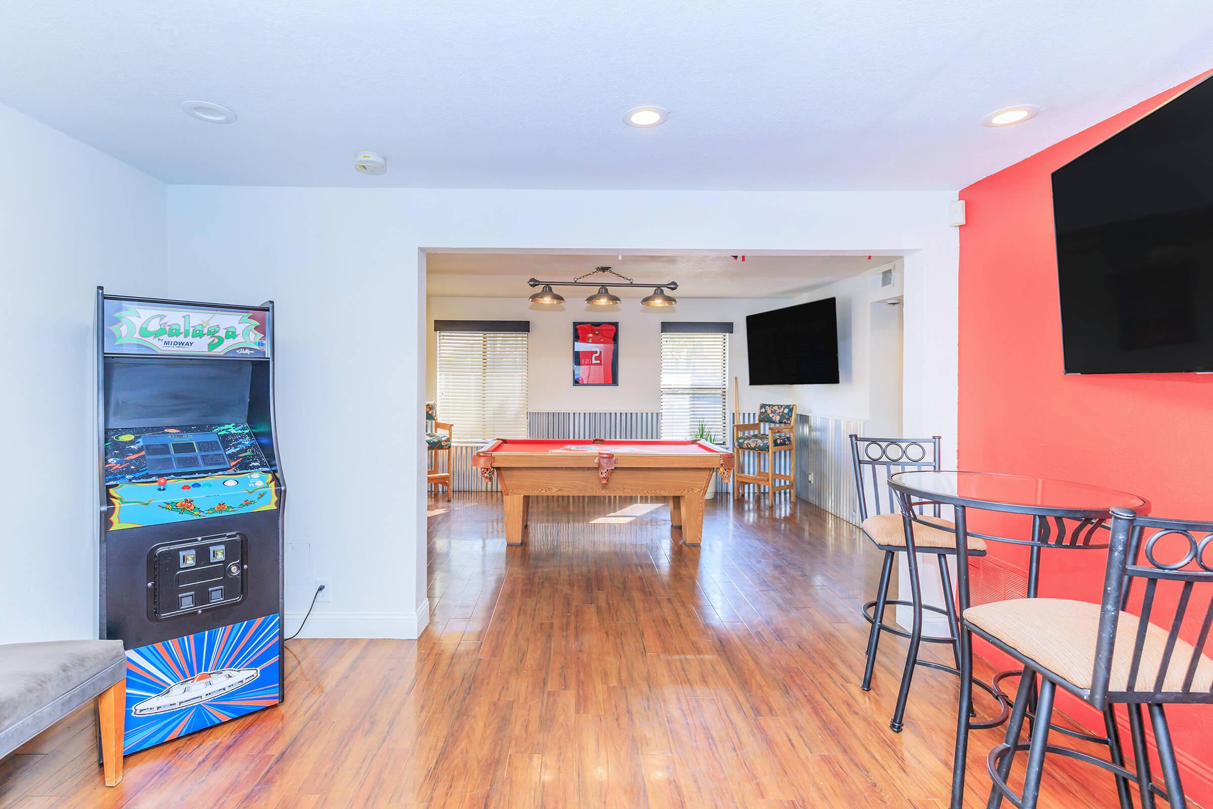 A bright and modern game room featuring a pool table, arcade game machine, and seating area. The walls are painted in a mix of white and orange, with two large flat-screen TVs mounted. Natural light floods the space through large windows, creating a cheerful atmosphere.