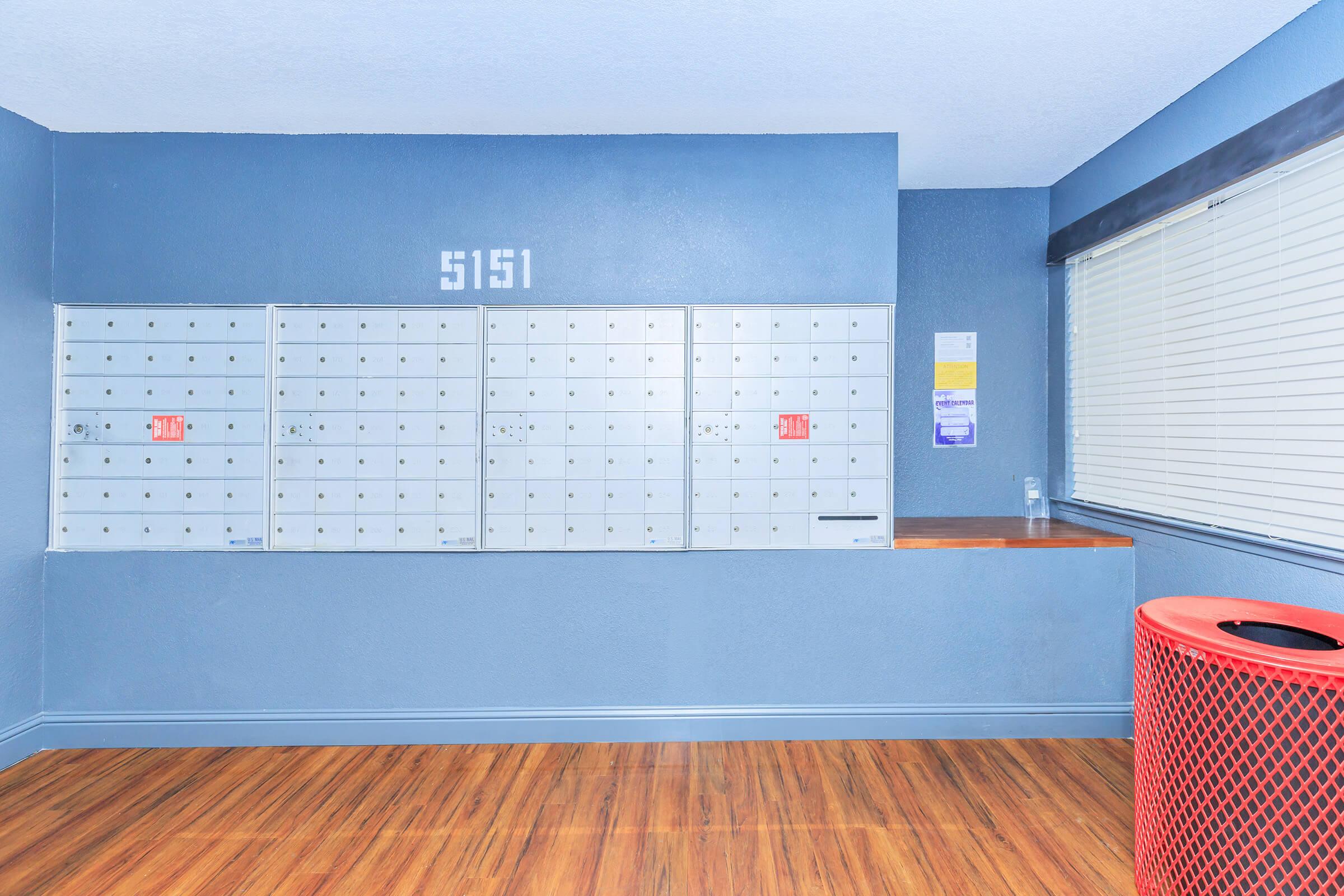 Row of locked mailboxes on a blue wall with the number "5151" displayed above. There is a counter area next to the mailboxes, and a red trash can is placed nearby on the wooden floor. A window with blinds lets in natural light, and there are informational papers on the wall.