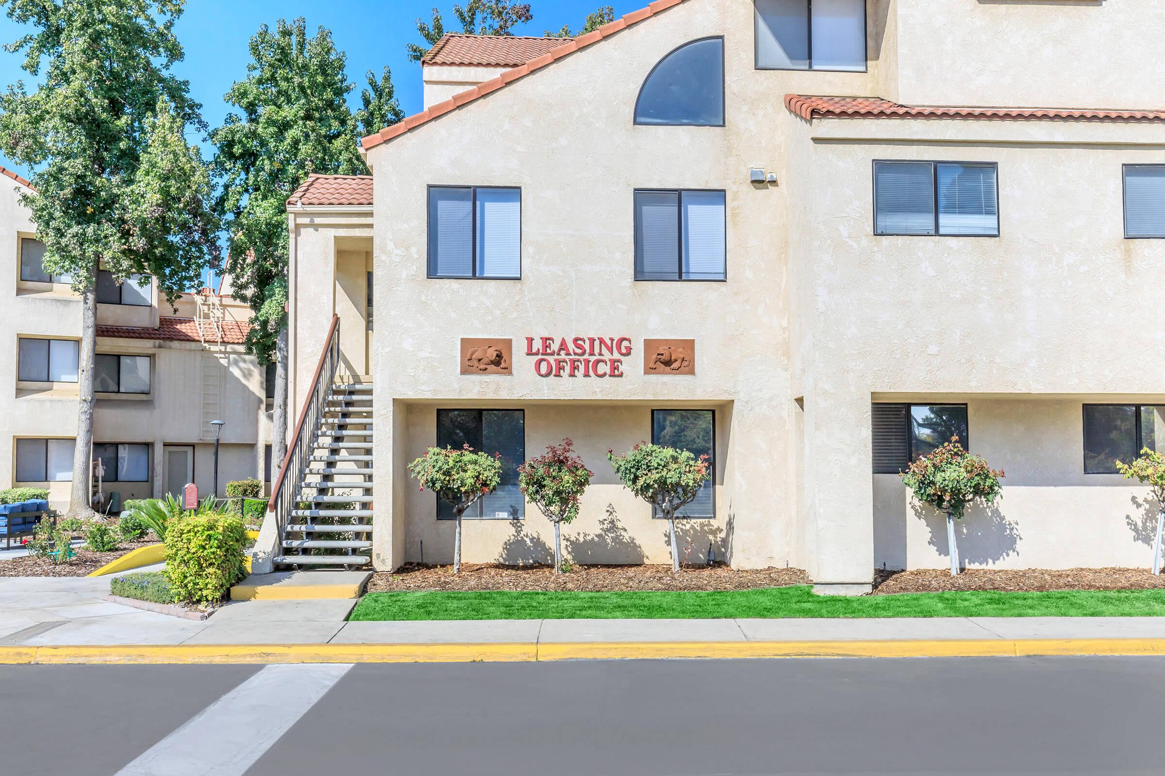 A leasing office building with a staircase leading to the entrance. The facade is light-colored with large windows and decorative elements. Lush green landscaping and bushes are situated in front, contributing to the inviting appearance of the office. Clear blue sky above.