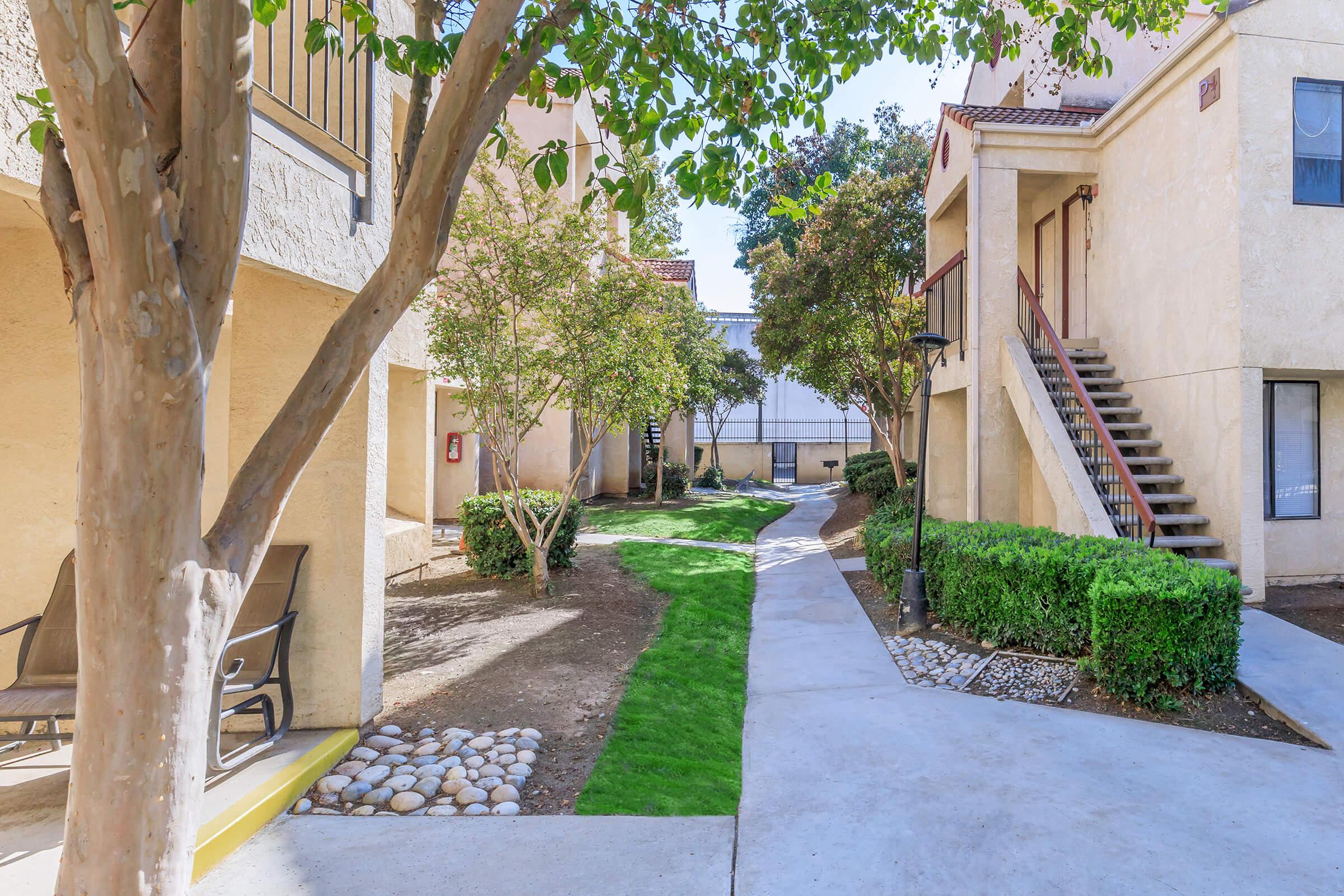 A walkway surrounded by two-story apartment buildings, with landscaped greenery, trees, and seating areas. The path features well-maintained grass and decorative gravel, leading toward stairways that access upper levels of the apartments. Clear blue skies are visible overhead.