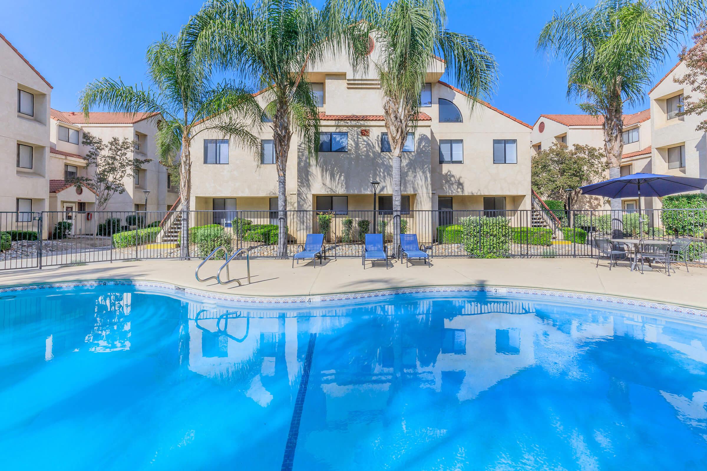 A clear blue swimming pool surrounded by lounge chairs and palm trees, with residential buildings in the background. The scene is sunny, showcasing manicured landscaping and a serene atmosphere ideal for relaxation.