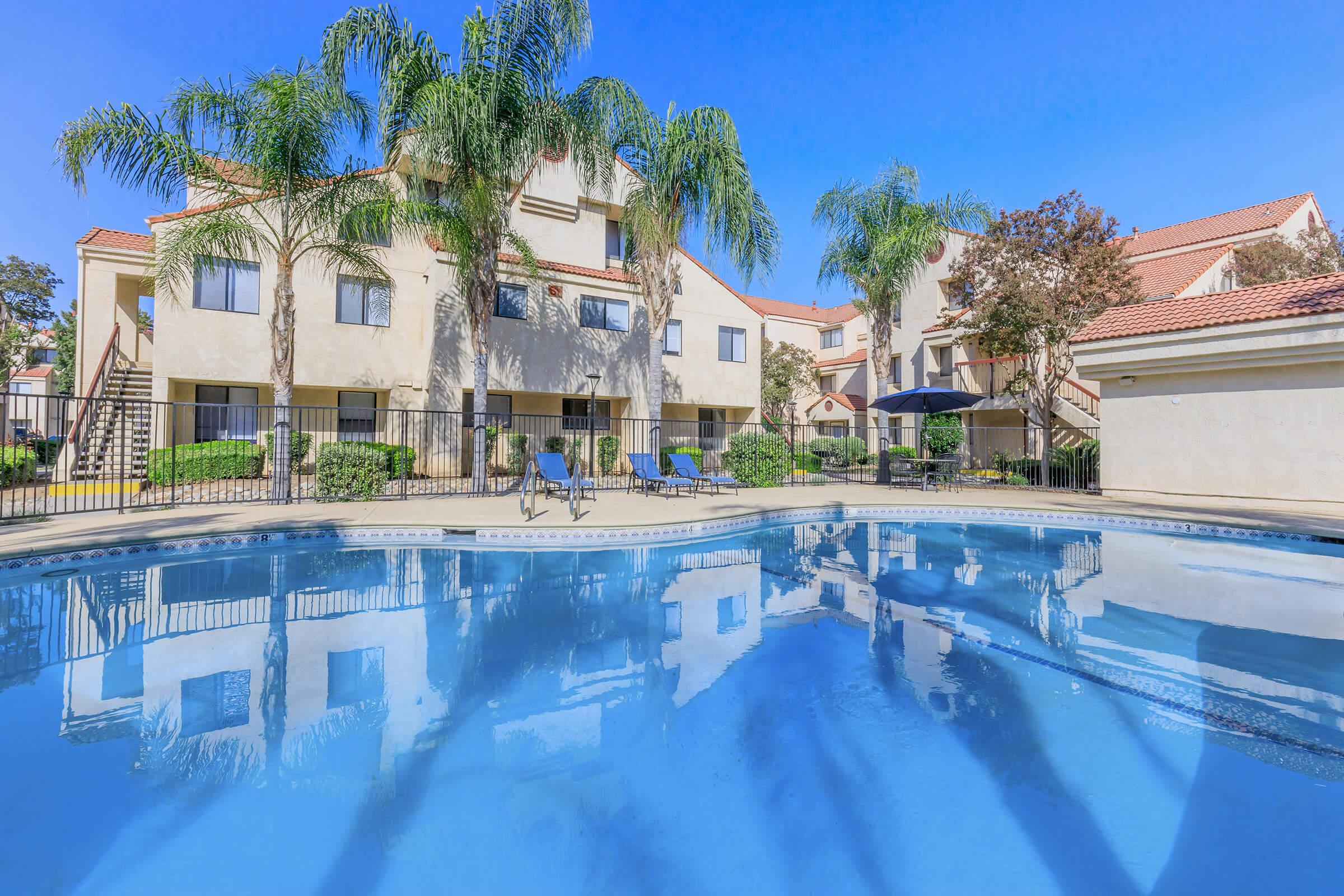 A tranquil swimming pool in the foreground, surrounded by palm trees and lush greenery. In the background, there are three-story apartment buildings with balconies. The sky is clear and blue, reflecting on the pool's surface, creating a serene and inviting atmosphere.