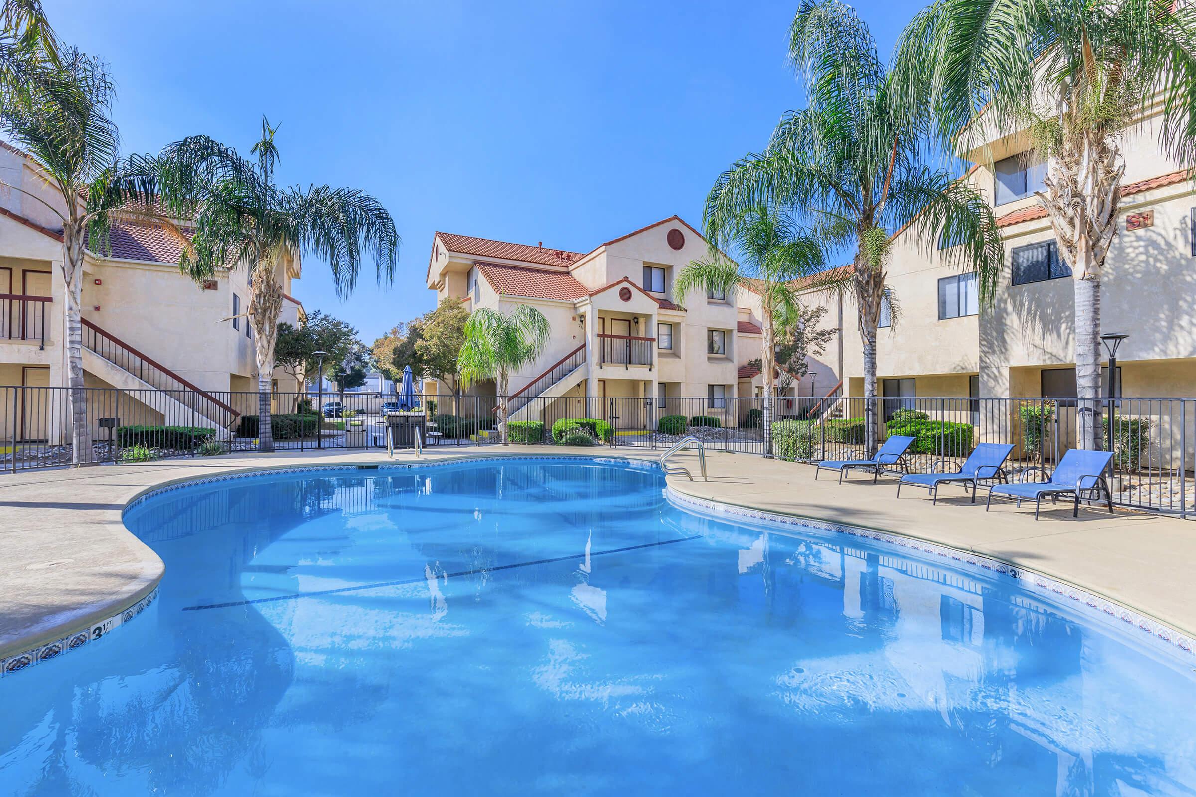 A clear blue swimming pool surrounded by palm trees and lounge chairs, with residential buildings in the background. The setting is bright and inviting, suggesting a warm and relaxing atmosphere typical of an apartment complex.