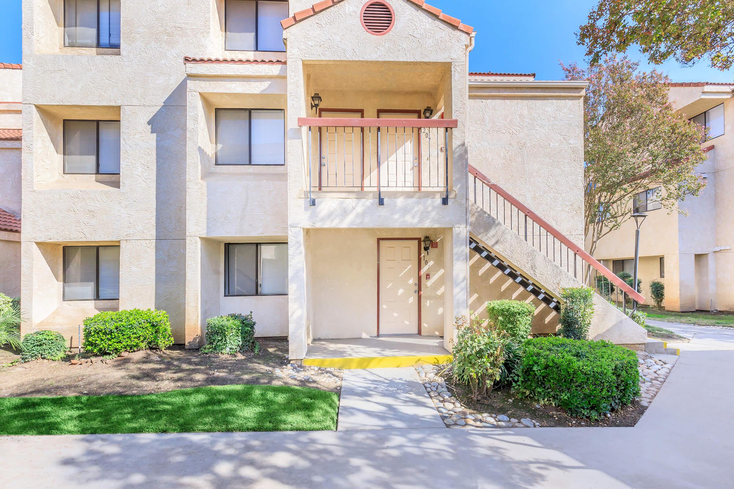 A three-story residential building with a balcony and stairs leading to the entrance. The exterior is light-colored with a textured finish. Surrounding greenery includes shrubs and small trees, and a walkway leads to the front door. The sky is clear and blue.