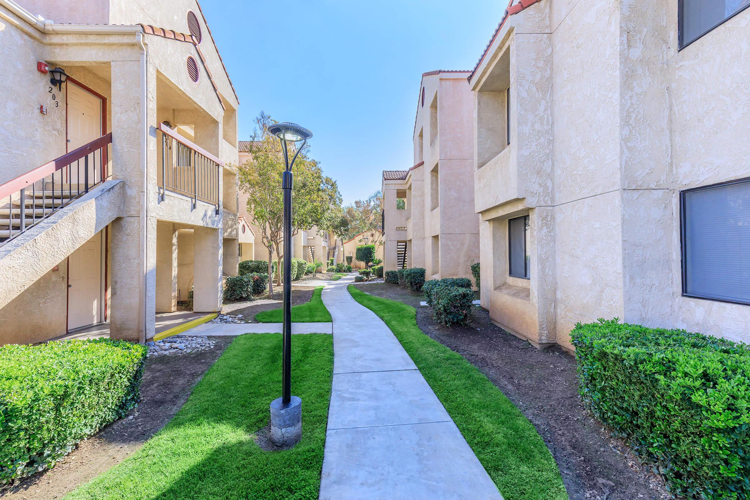 A well-maintained pathway leading through an apartment complex, flanked by neatly trimmed hedges and small trees. The buildings, with light-colored exteriors, are set against a clear blue sky, creating a tidy and inviting atmosphere. A lamppost adds to the charm of the walkable area.